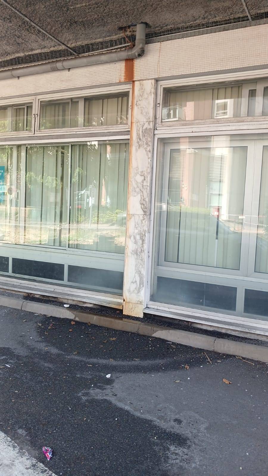 Exterior storefront and ground floor office film location in Watford WD18 available via Love Locations, featuring weathered modernist architecture, large glass display windows with vertical blinds, a marble-effect support pillar, and a concrete overhang.