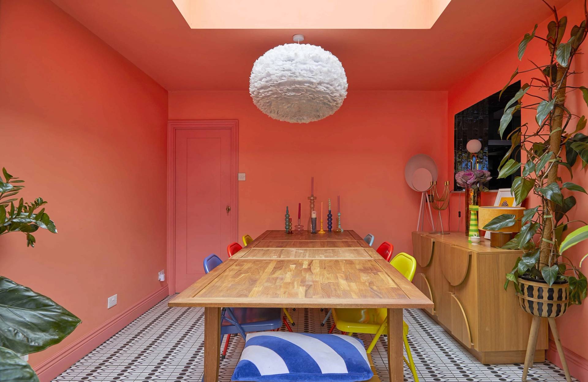 Spacious coral orange dining room with a long wooden table, yellow and red chairs, and a white feather pendant light, Love Locations Catford.