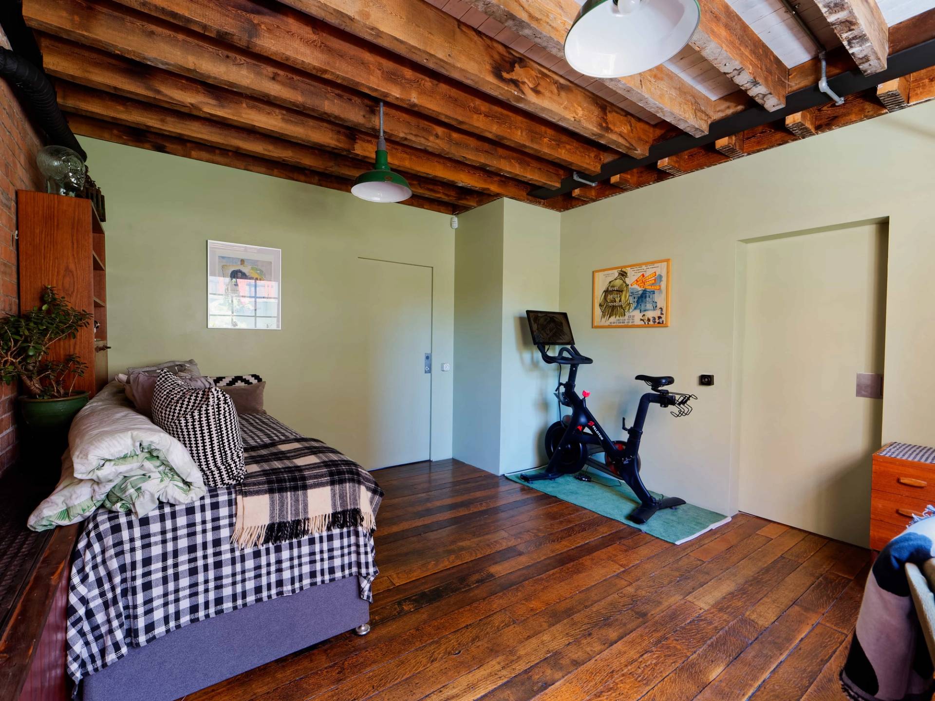 Private home gym corner featuring a Peloton exercise bike, exposed timber ceiling, and plaid sofa bed, Love Locations E14.