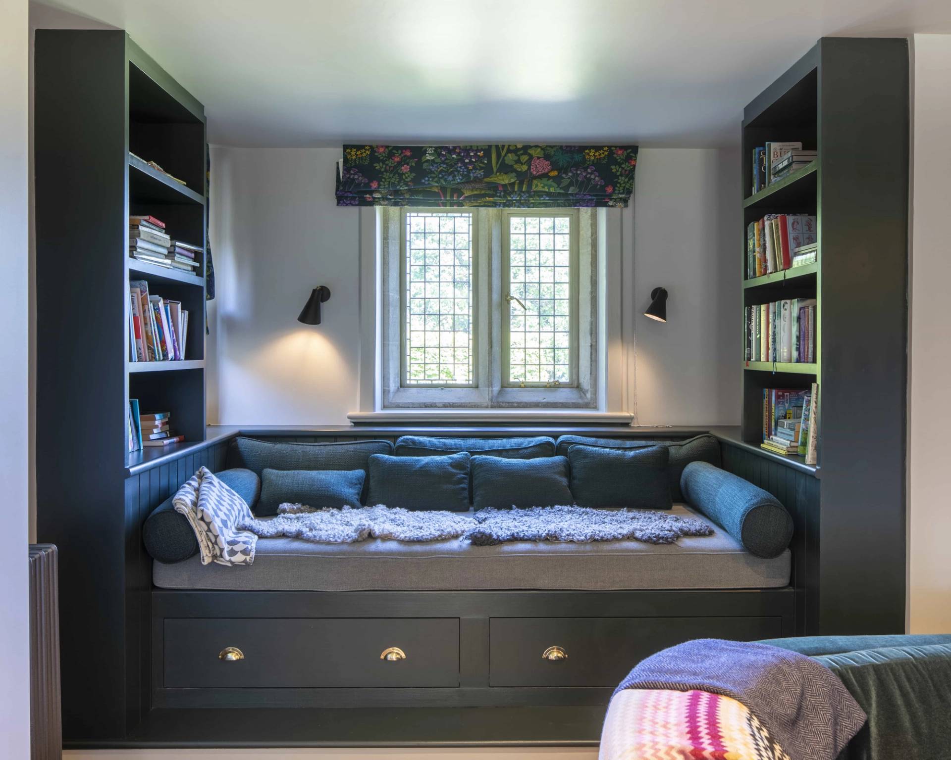 Cozy window nook featuring a dark built-in upholstered daybed set between bookcases, Love Locations OX7.