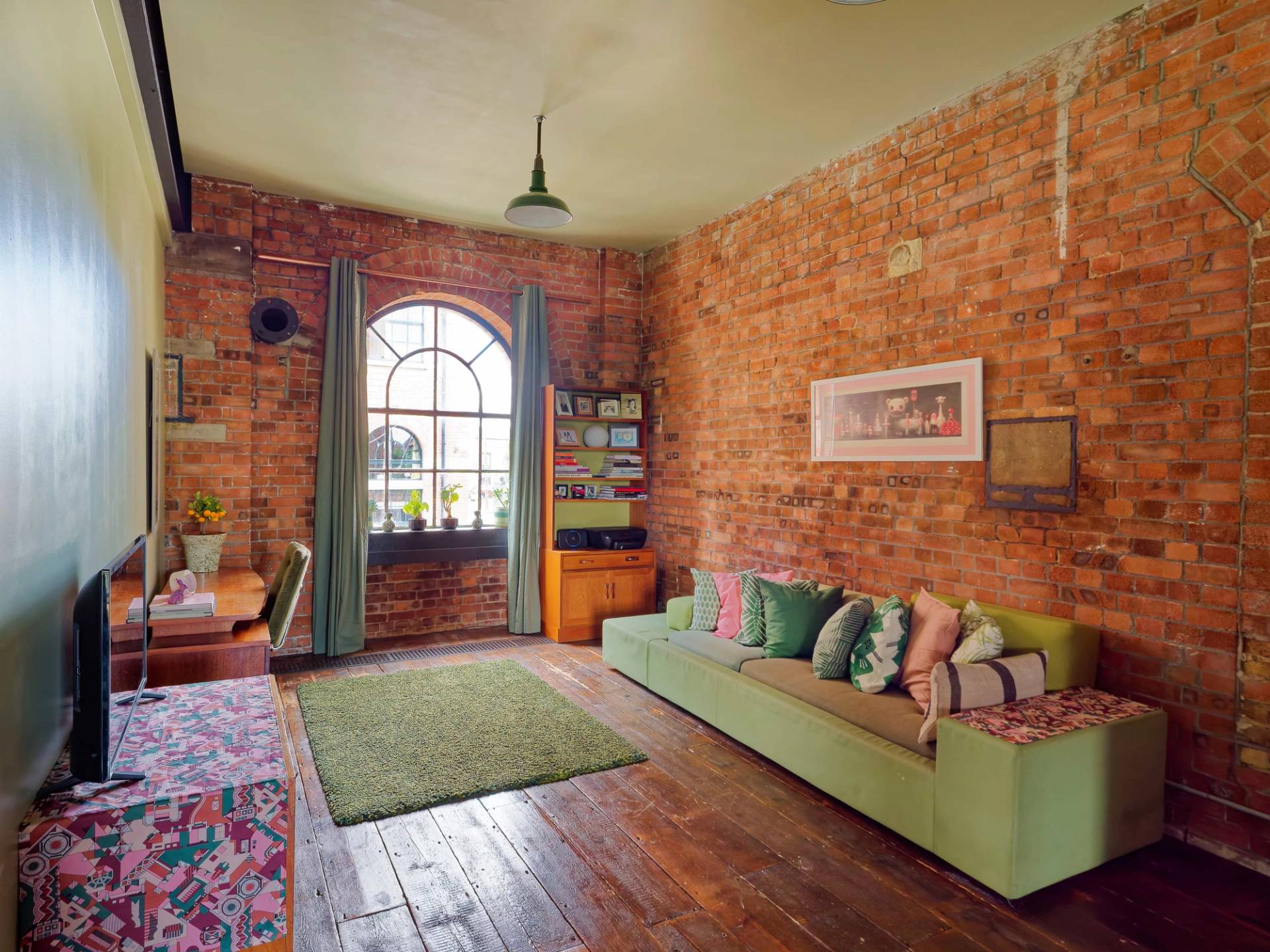 Industrial loft room with arched warehouse window, exposed red brickwork, green bench sofa, and vintage desk, Love Locations E14.