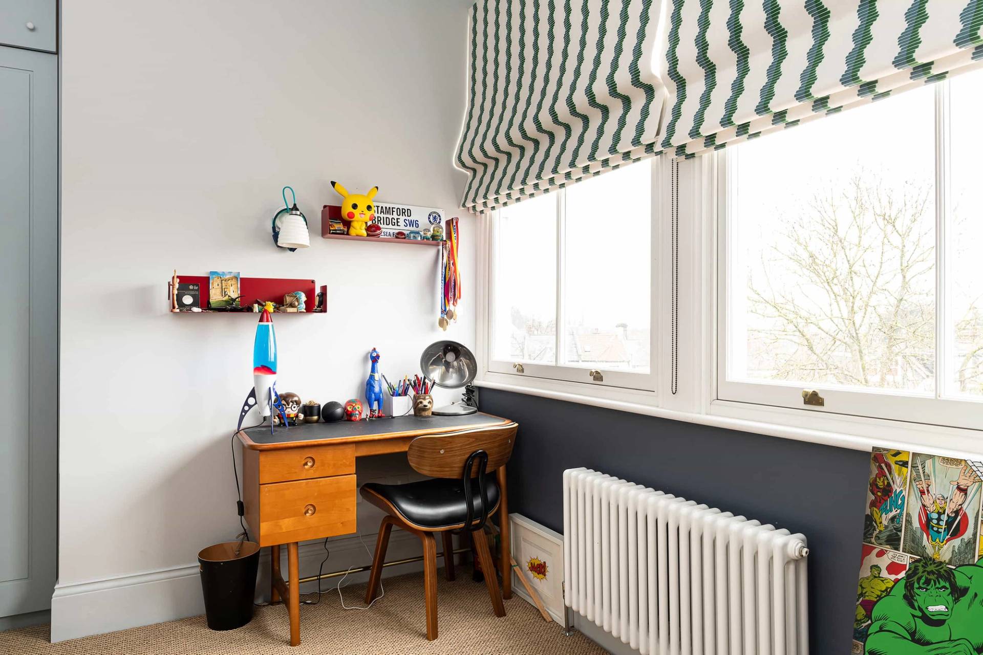 Children's study desk corner with dark blue lower walls, wooden desk, and green striped window blinds, Love Locations.