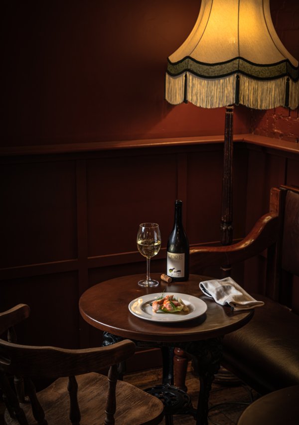 Elegant food and wine editorial display showcasing a plated dish, a bottle of white wine, and a filled wine glass under a fringed vintage lamp, ideal for menu marketing, Love Locations.