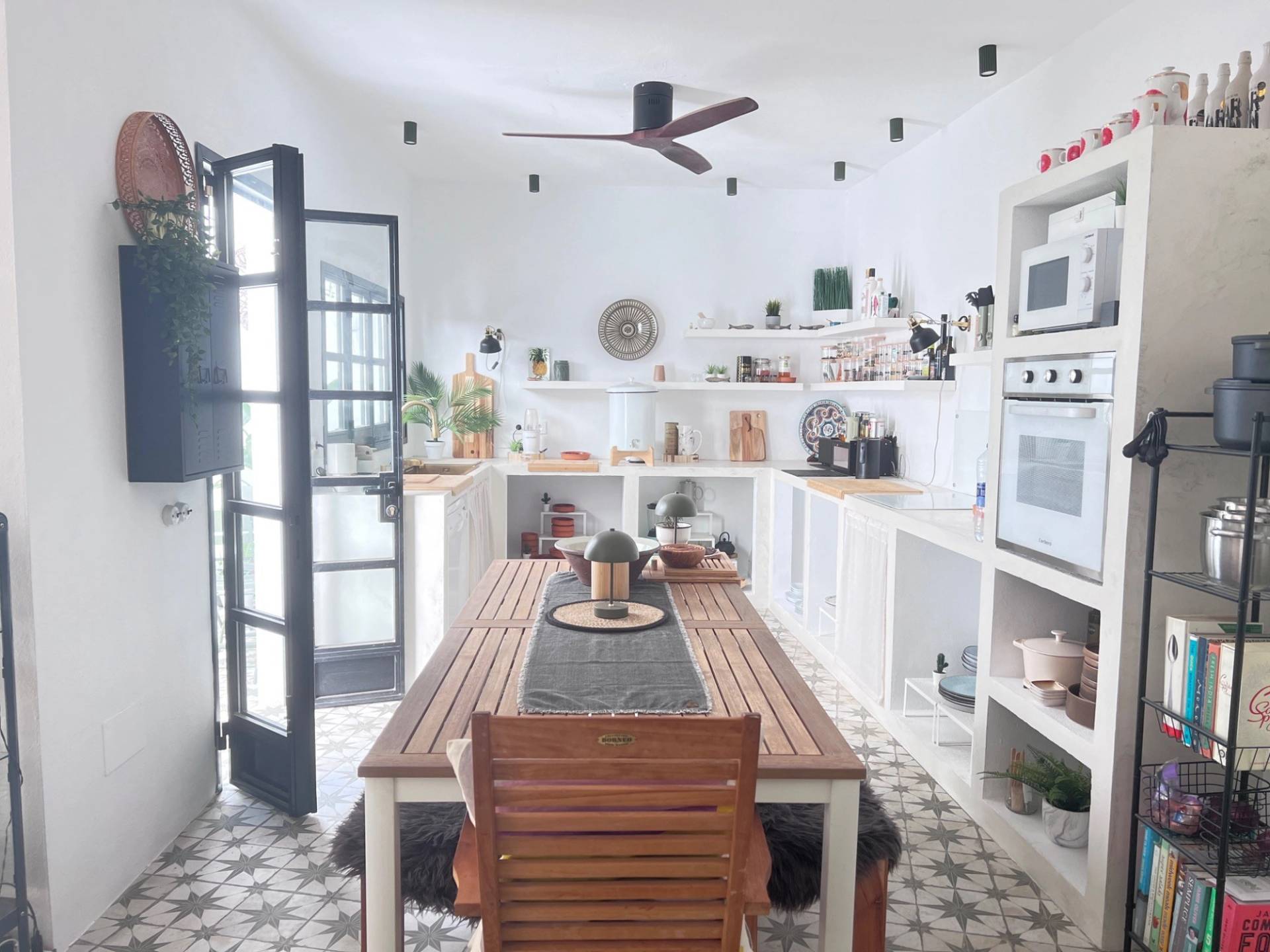 Rustic Spanish kitchen shoot location in Jimena de la Frontera via Love Locations, featuring open wood shelving, a long dining table, built-in shelving, and dark framed patio doors.
