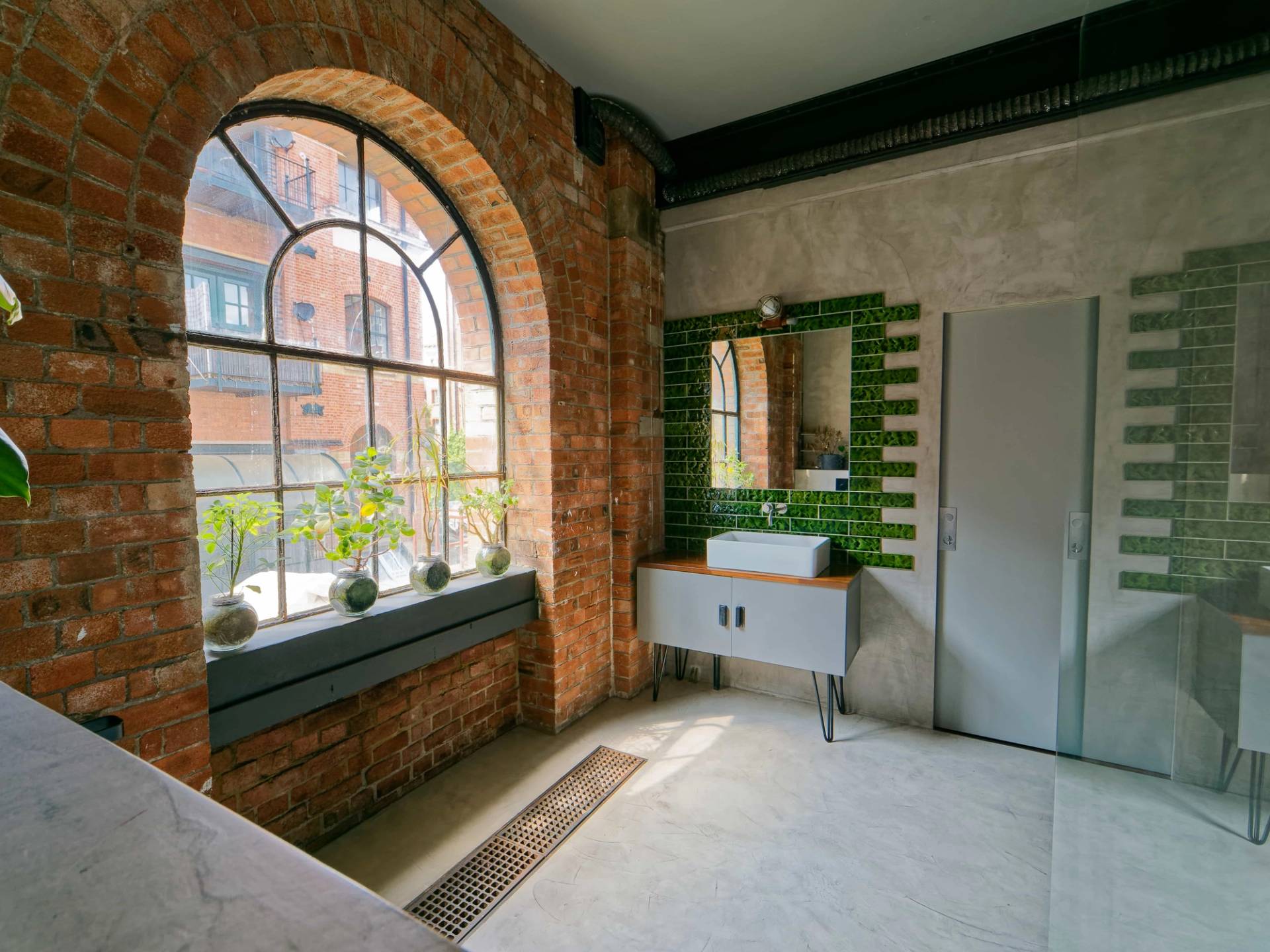 Micro-cement bathroom with green tiled vanity backsplash, white sink console, and arched warehouse window views, Love Locations E14.