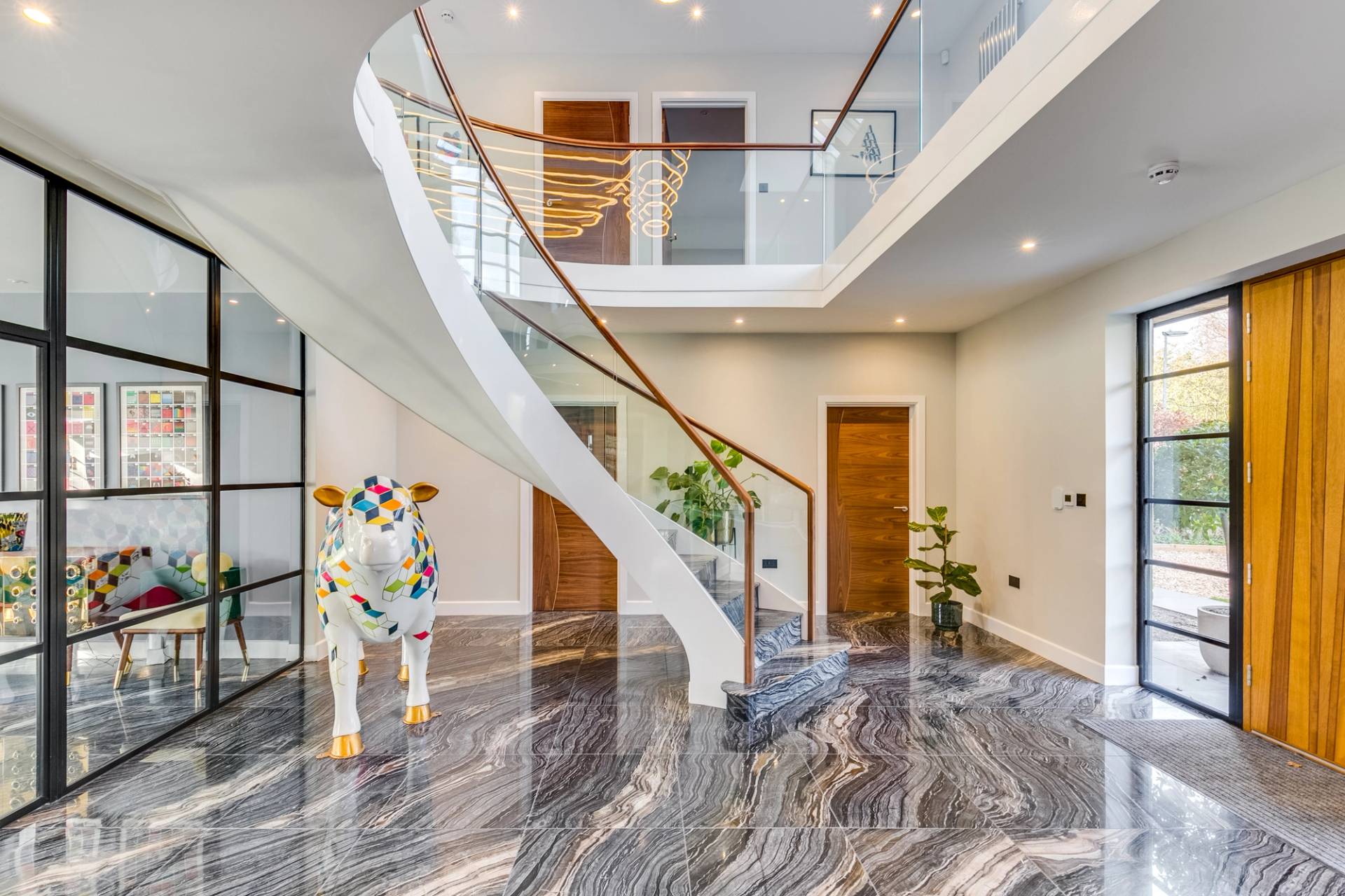 Grand double-height entrance hall featuring a sweeping curved staircase, marble floors, and life-size painted cow sculpture, Love Locations CB2.