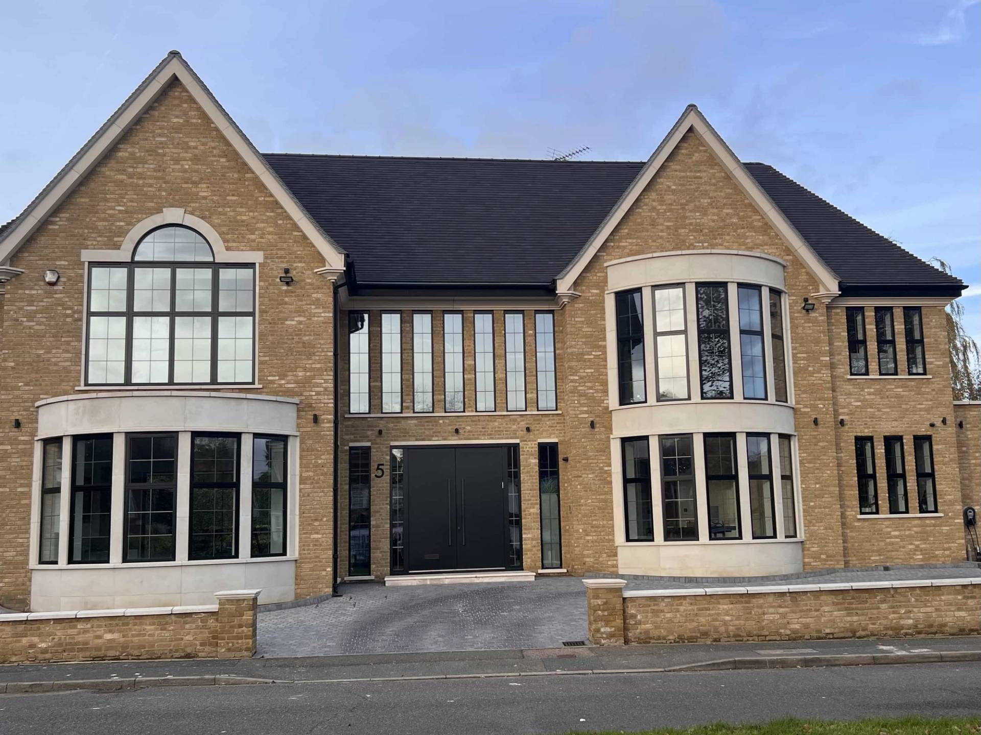 Front exterior facade establishing shot of a modern detached yellow-brick mansion in Finchley Church End, N3 featuring bay windows, black double entrance doors, and paved driveway, the ultimate hero backdrop for luxury car commercials, TV drama establishing shots, and commercial film productions via Love Locations.