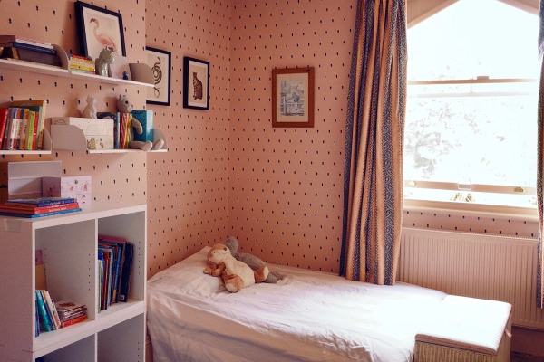 Charming children's single bedroom with dotted pink wallpaper, a wooden bed frame, and classic window curtains, Love Locations.