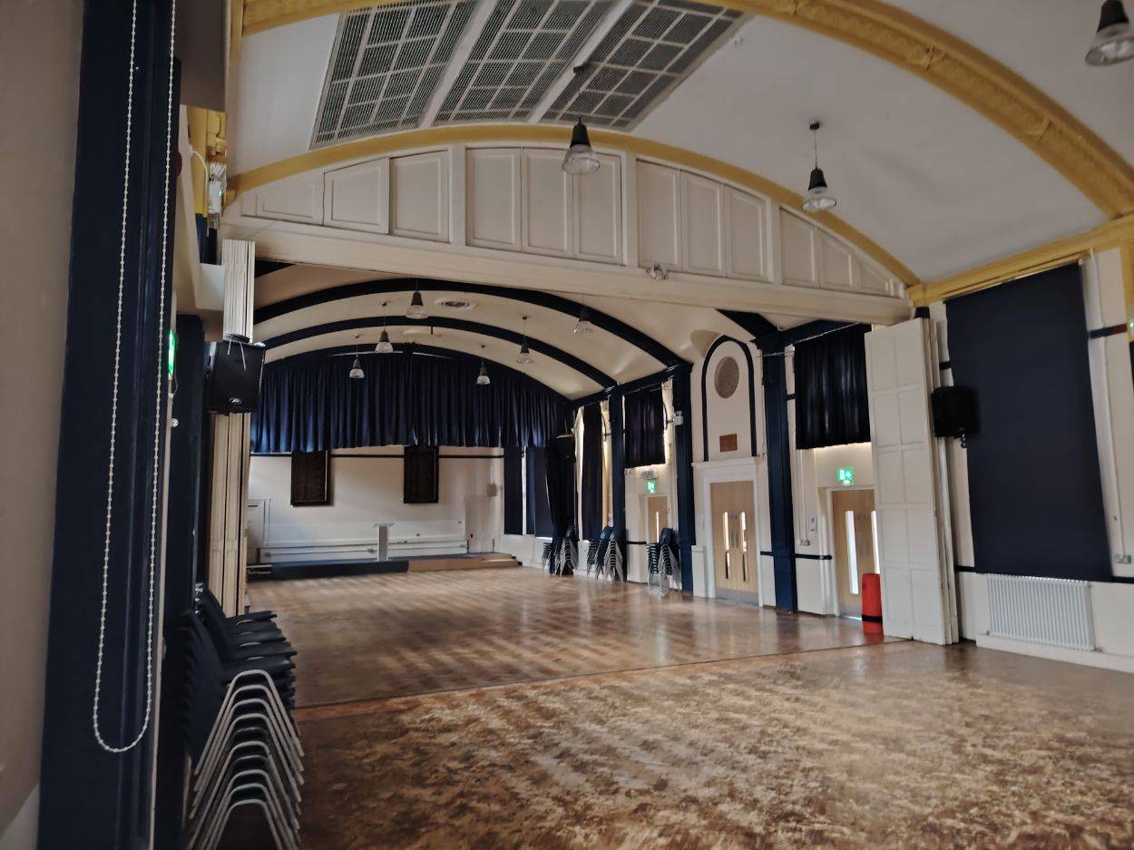 Wide view of an empty school assembly hall photo location showing polished parquet parquet flooring, vaulted ceiling trusses, and blue stage curtains by Love Locations.