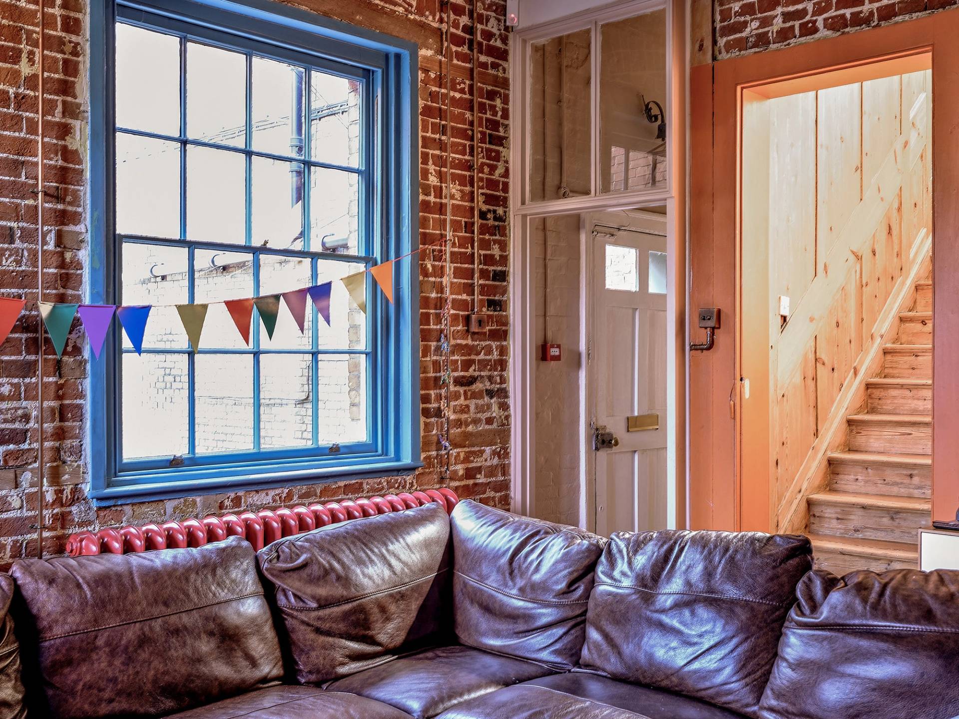 Exposed red brick wall living room with blue framed windows, leather sofa, and bunting decorations, Love Locations ME13.