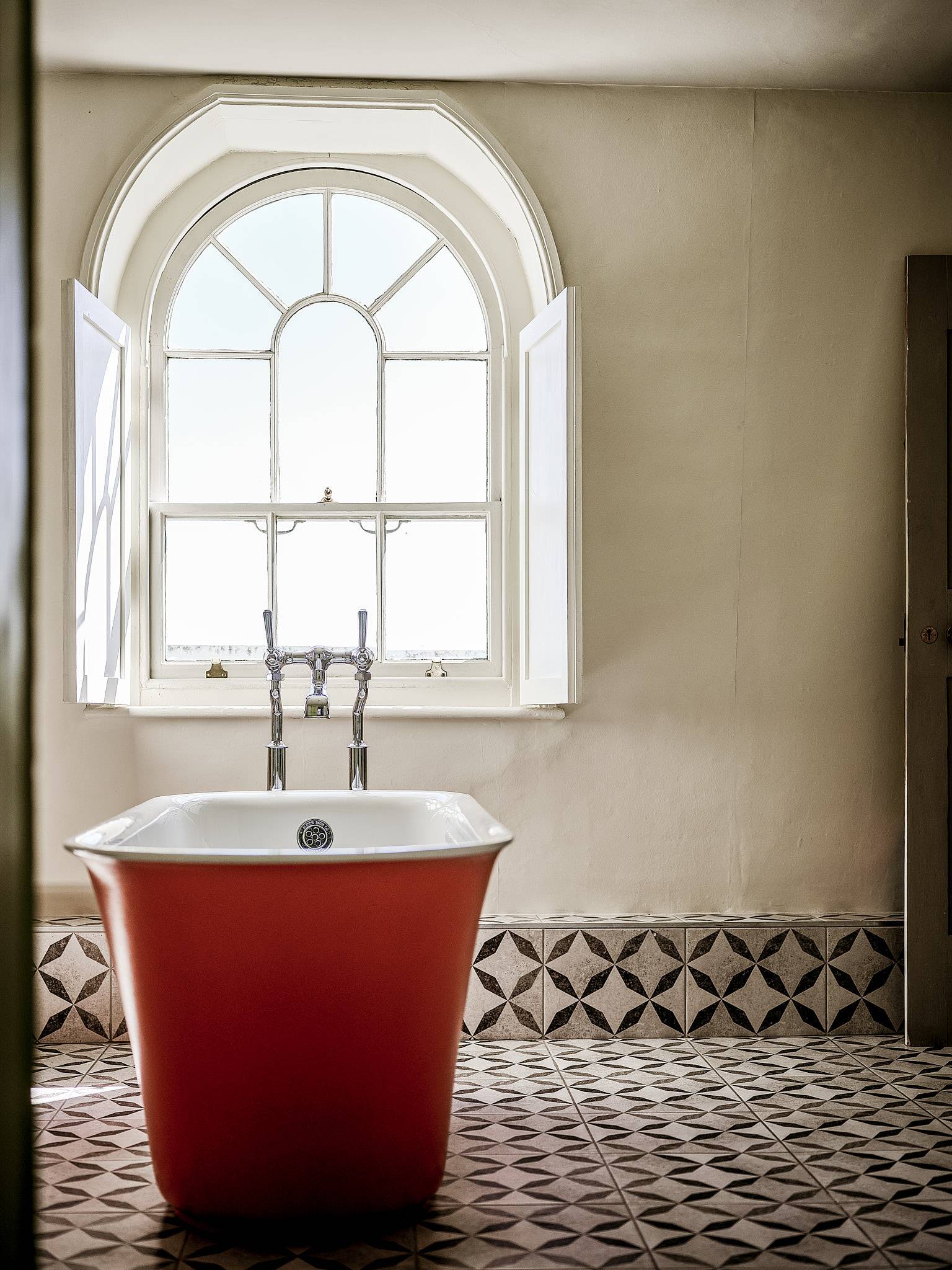 Terracotta red soaking tub set on patterned geometric floor tiles before an arched sash window, ideal for wellness brand shoots, Love Locations ME13.