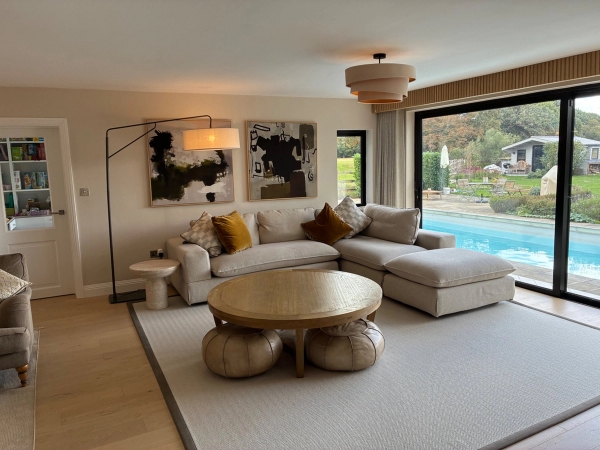 Spacious open-plan living room location house in Essex CM4 from Love Locations, featuring modern grey sectional sofas, a natural round wooden coffee table, and large sliding glass doors leading to the garden pool.