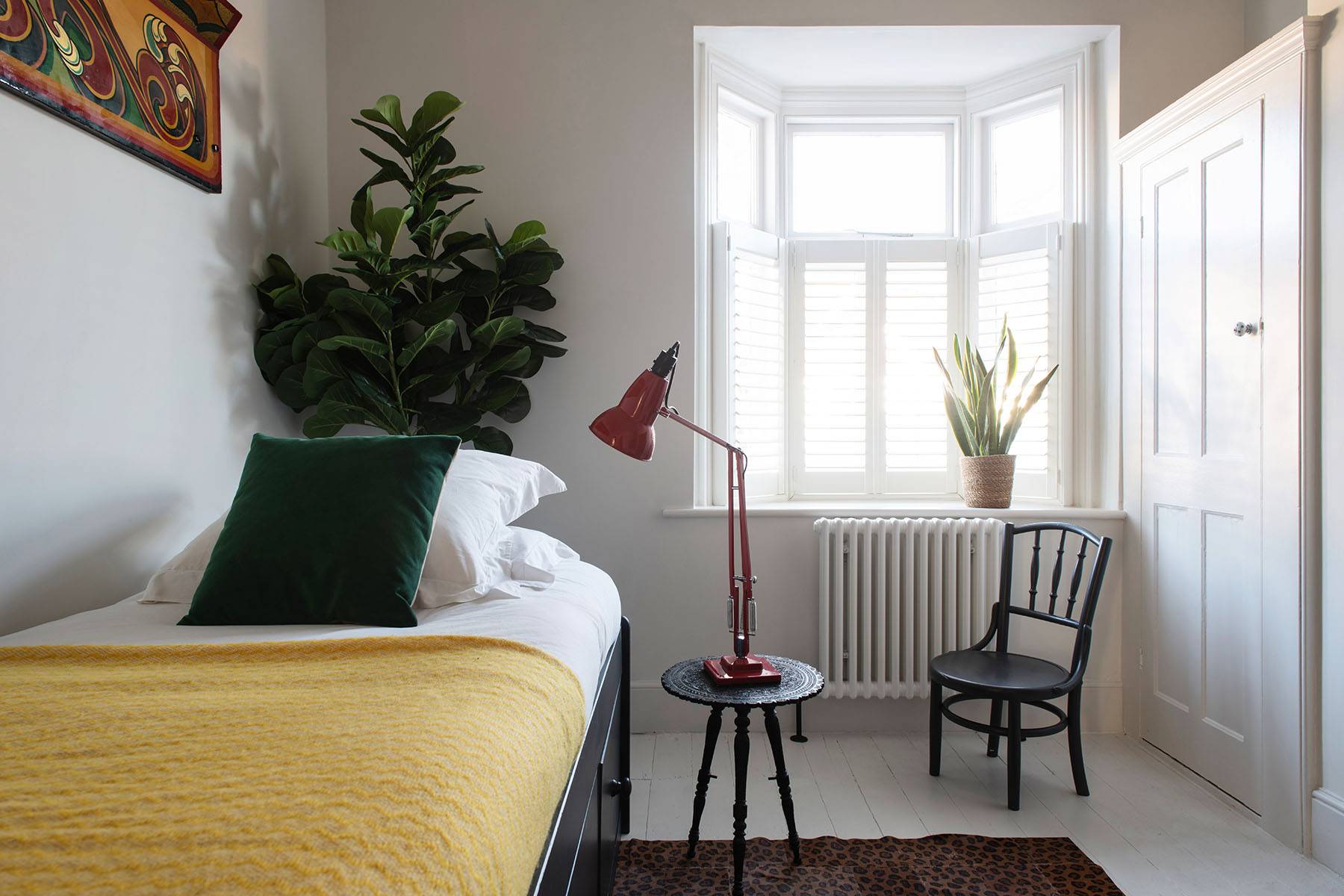 Light-filled bedroom corner with red anglepoise lamp, window shutters, and green velvet accent cushion, Love Locations.