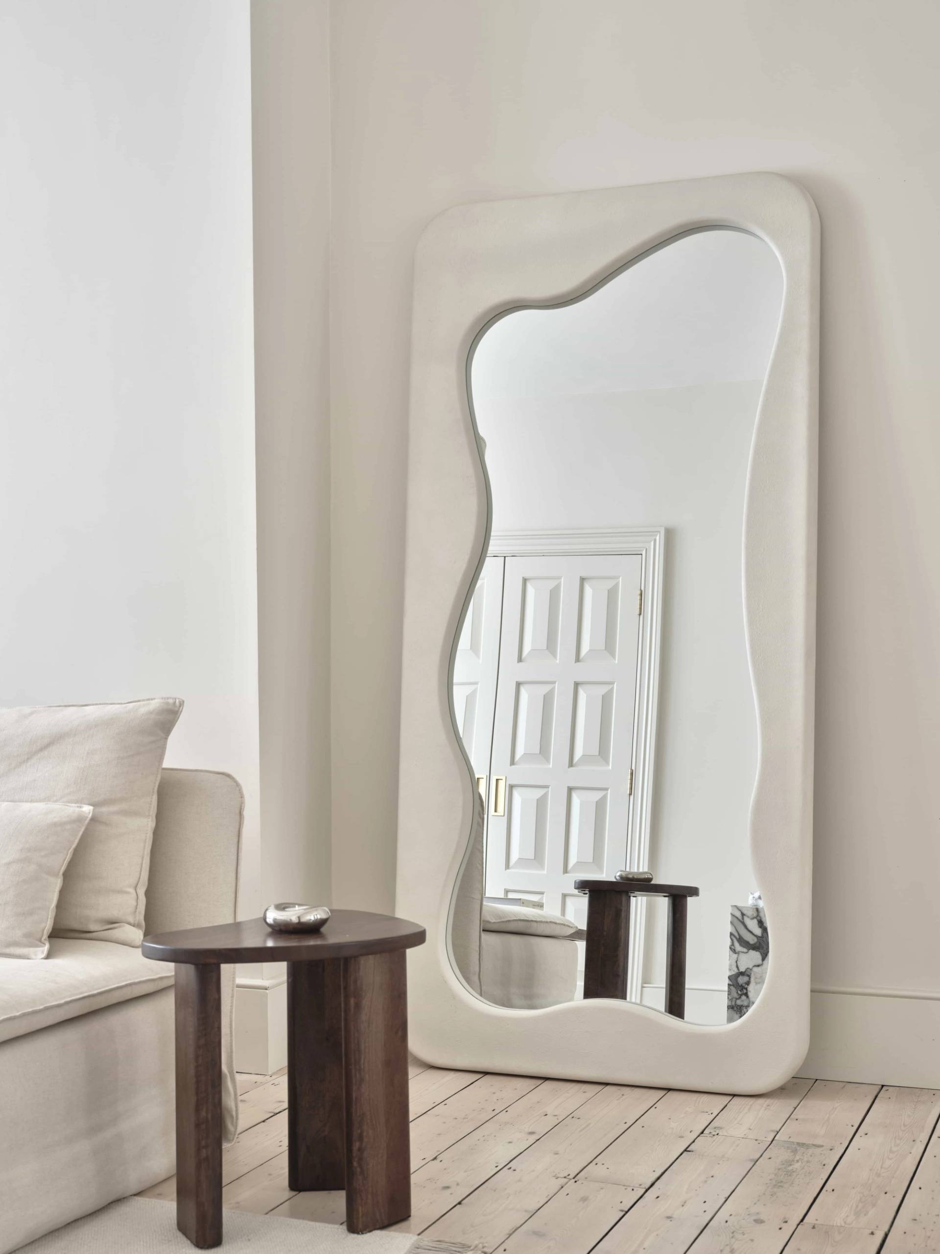 Wavy full-length floor mirror standing beside a dark wood side table in a neutral living room, Love Locations E4.