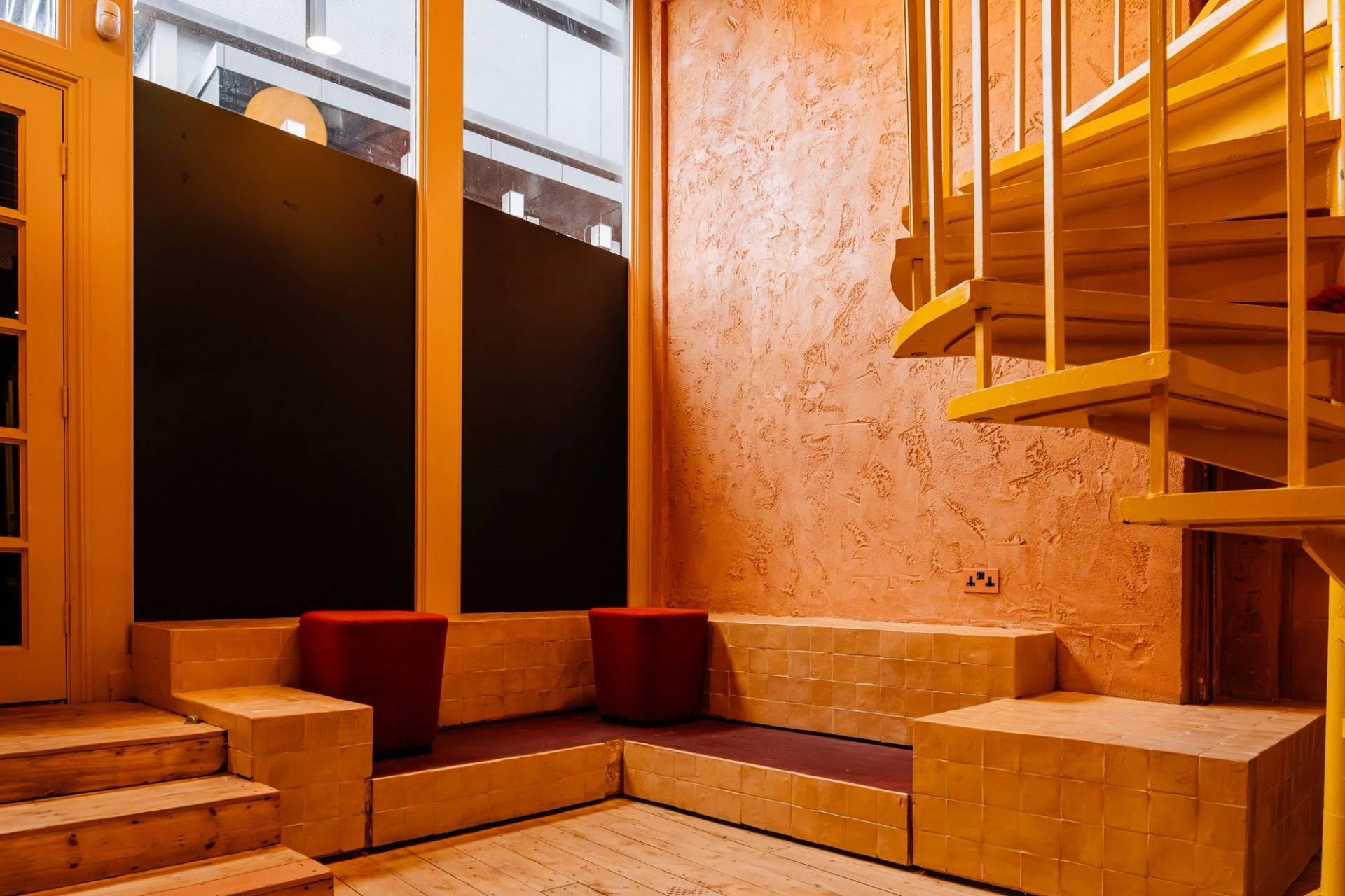 Cozy lounge nook showcasing rich terracotta plaster finish, yellow architectural stairs, and plush red cushions by Love Locations.