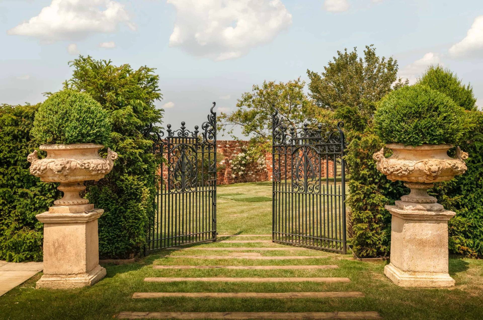 Wrought iron garden gates opening onto manicured lawn pathways and stone urn planters in Gloucestershire GL51 from Love Locations.
