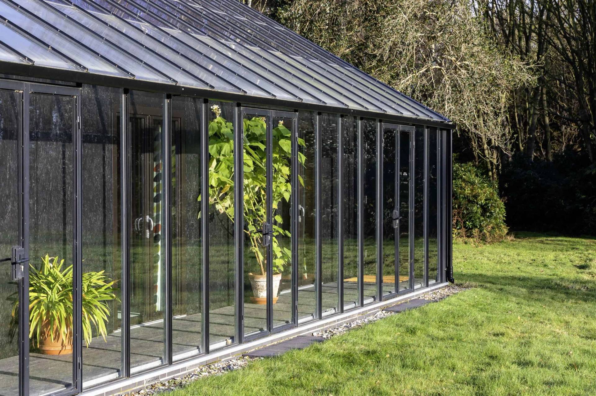 Sunlit glass greenhouse gallery corridor with black metal frame and stone gravel base, Love Locations IP17.