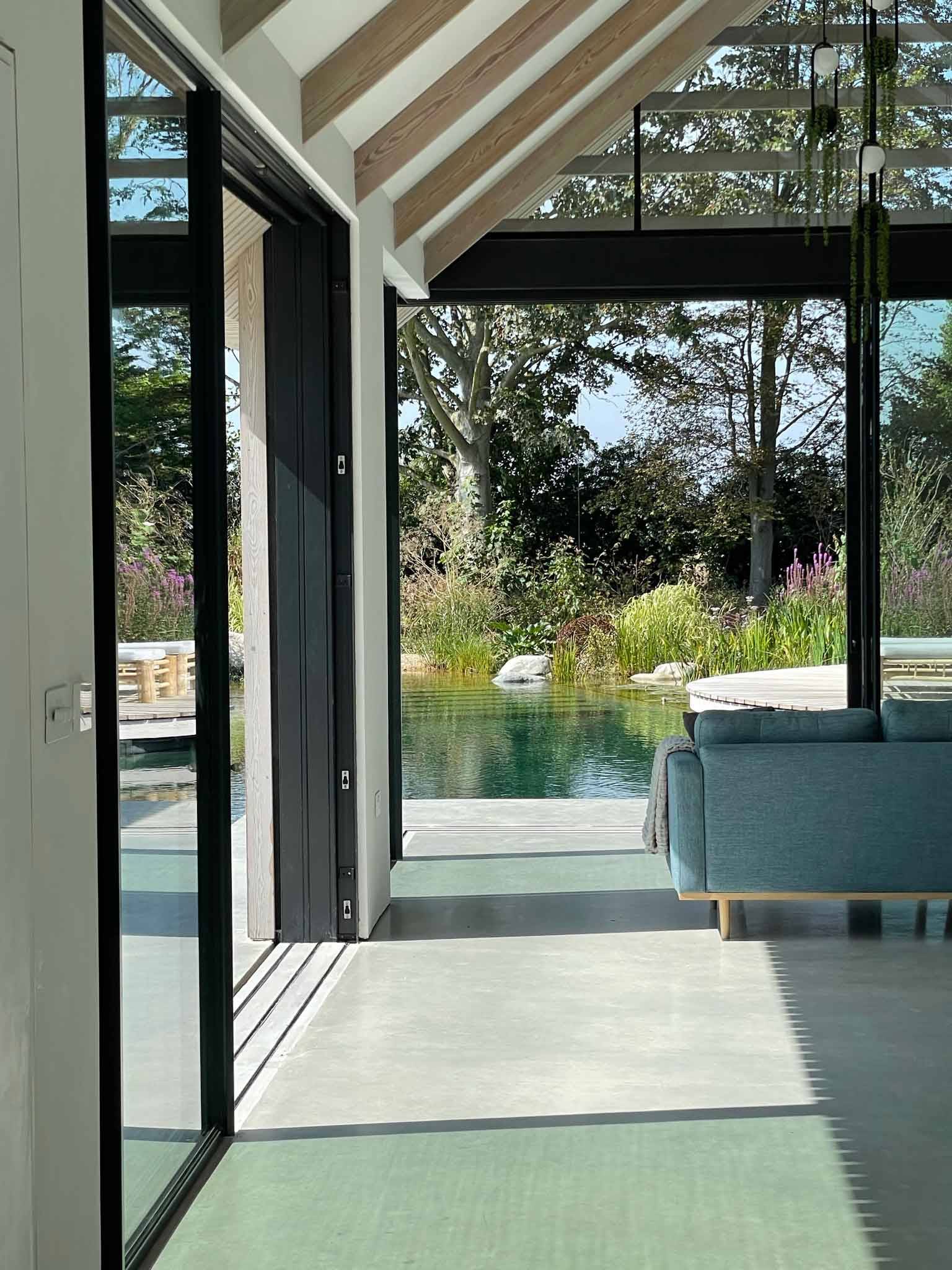View looking out from an open-plan lounge through floor-to-ceiling sliding glass doors onto a natural pool, perfect for indoor-outdoor commercial shoots, Love Locations PO18.