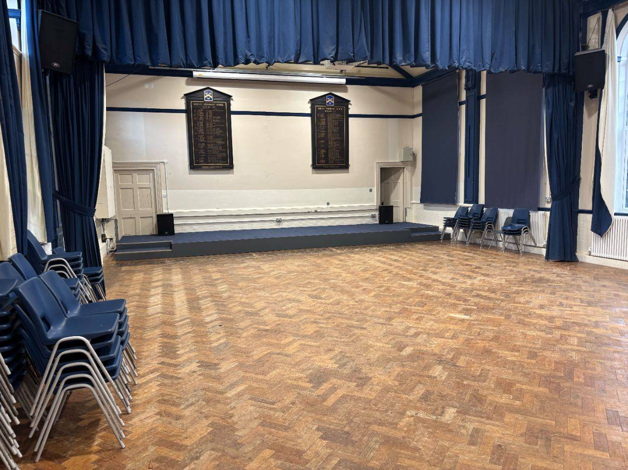 Traditional school performance hall filming space featuring a wooden parquet floor, elevated stage, and blue window drapes through Love Locations.