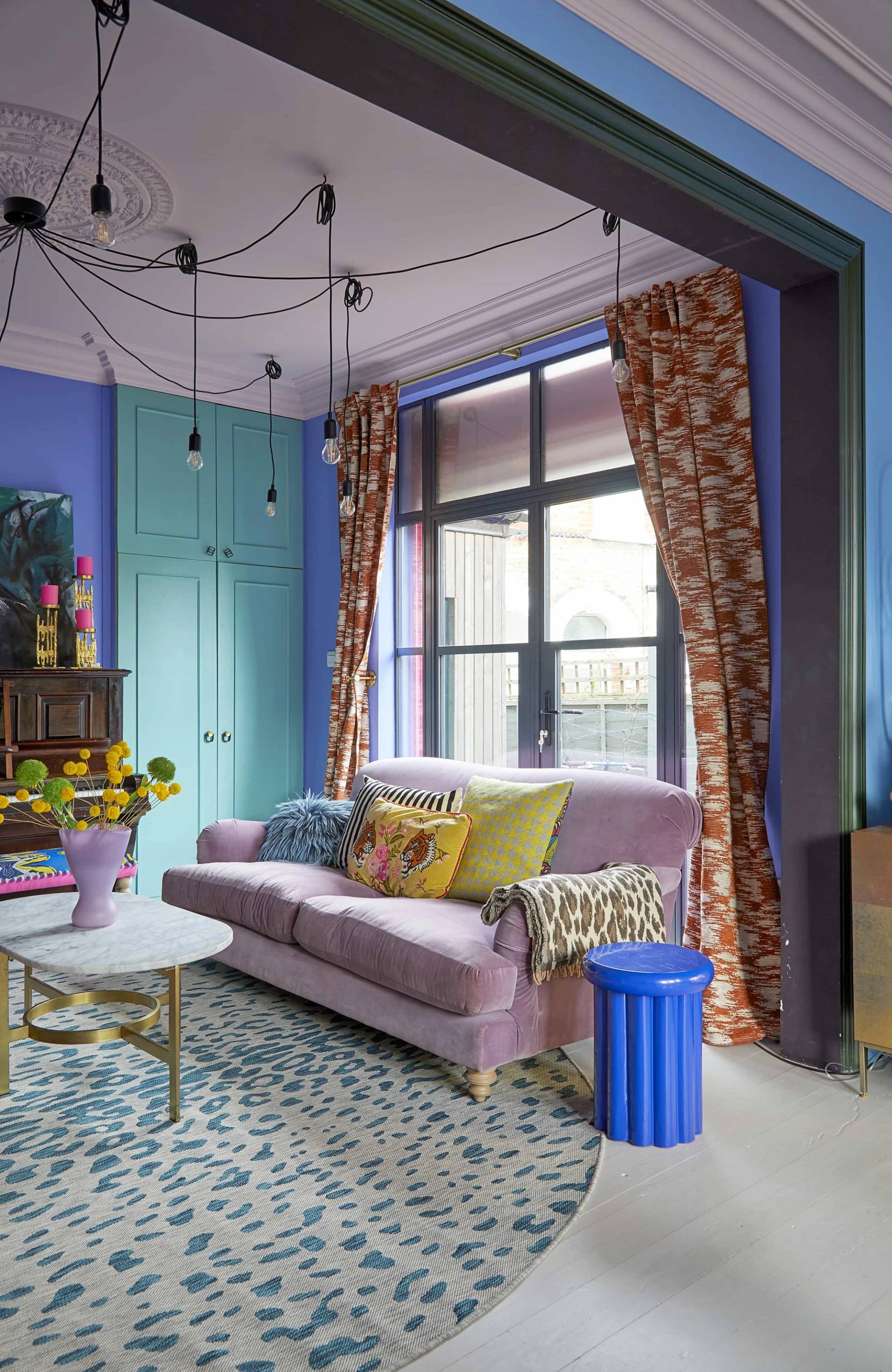 Spacious lounge area with a plush lilac sofa, blue side table, patterned curtains, and vibrant blue walls, Love Locations Catford.