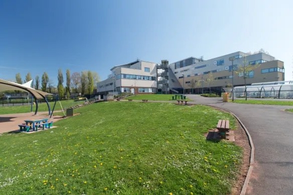 Love Locations contemporary school building exterior Bristol: Looking across a pristine green lawn and picnic benches toward a modern multi-story school campus facade, ideal for institutional establishing shots.