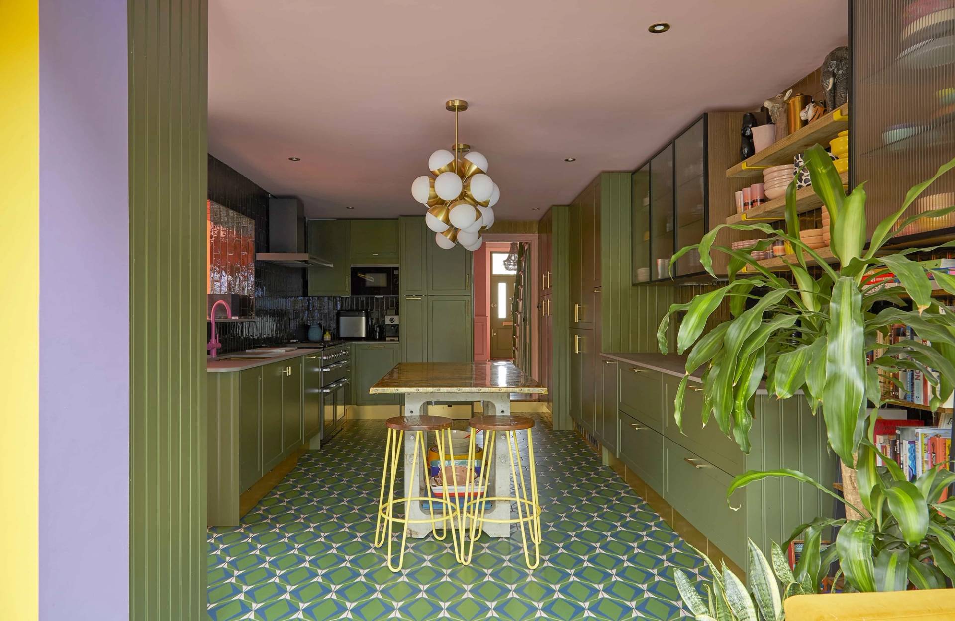 Symmetrical shot of an olive green kitchen with green patterned tiles, brass hardware, and a mobile island, Love Locations Catford.