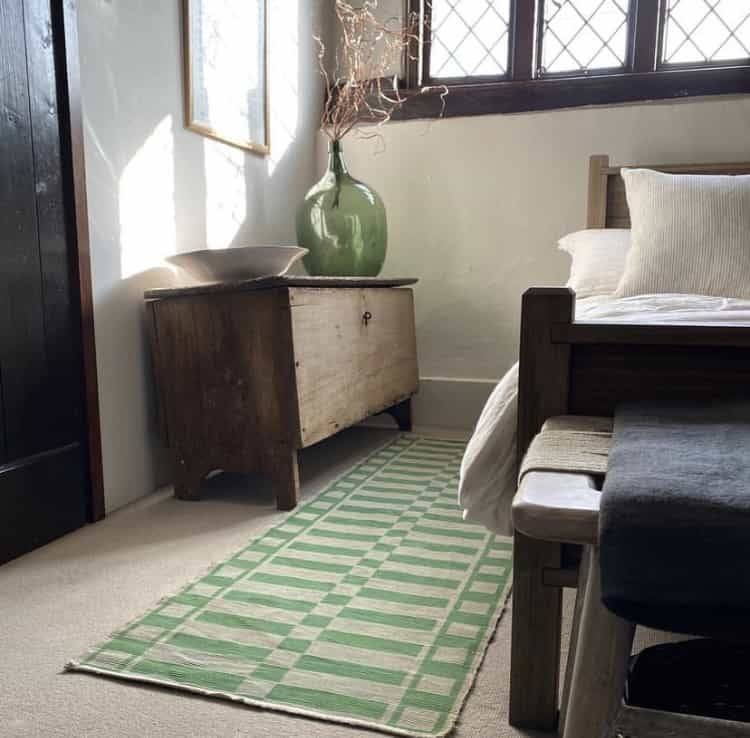 Historic cottage bedroom corner featuring a wooden chest, a green patterned rug, and crisp white bedding under low timber ceilings, Love Locations.