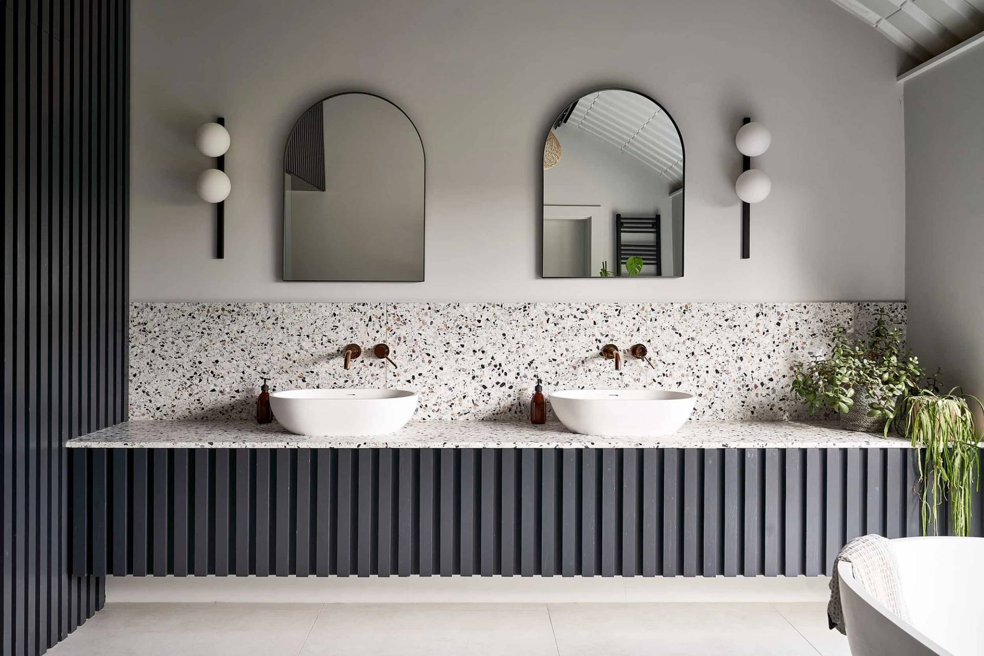 Double vanity bathroom unit featuring twin white vessel sinks, dark fluted panelling, terrazzo wall tiles, and arched mirrors, Love Locations IP30.