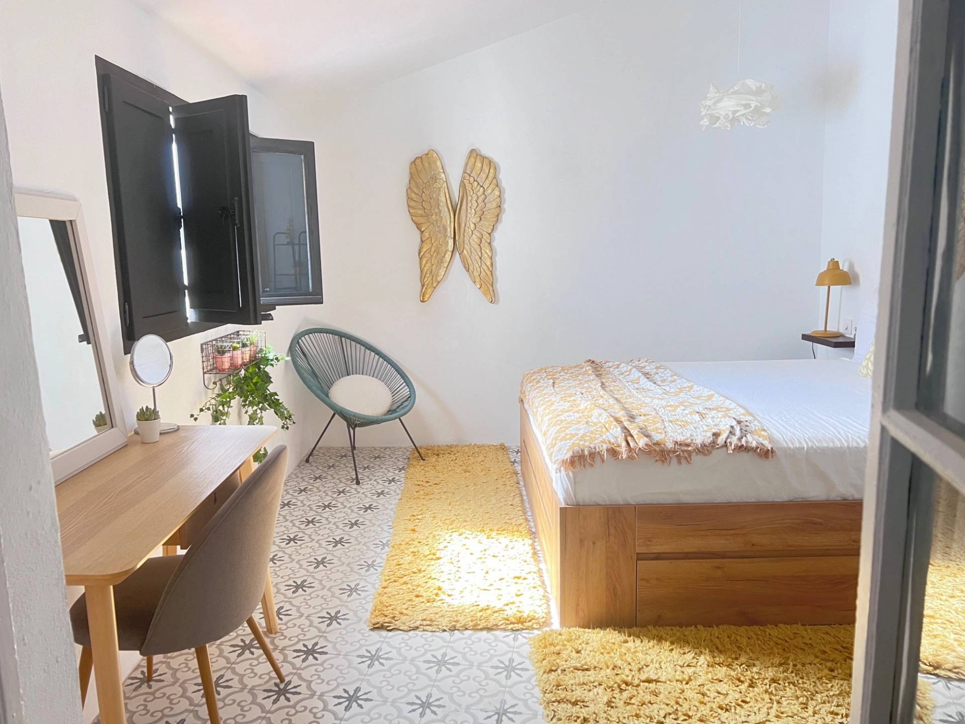 Sunlit guest bedroom film location in Jimena de la Frontera via Love Locations, showcasing a wooden bed frame, mustard yellow throw rug, and patterned tiled flooring.