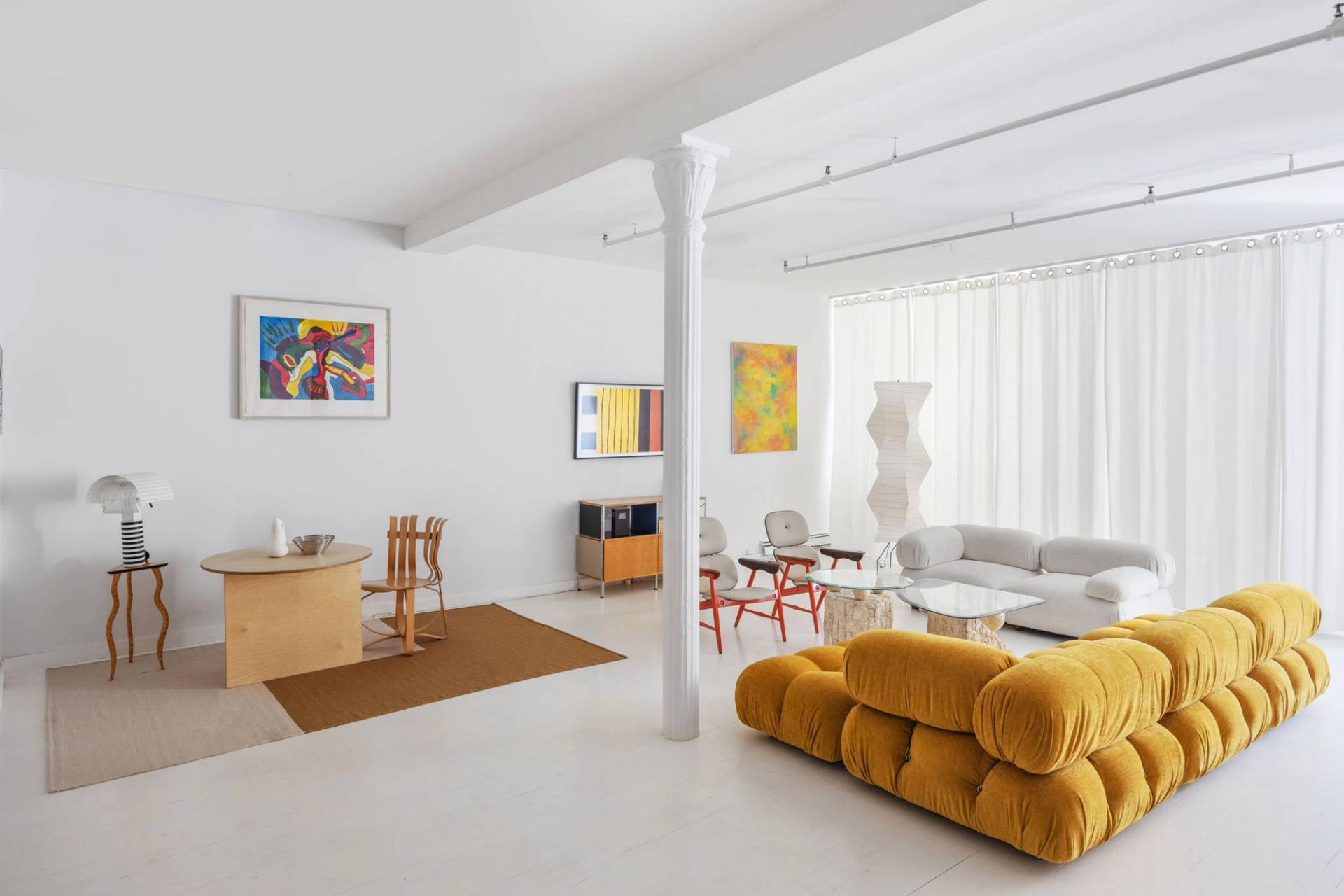 Sprawling all-white artist loft film location in Tribeca NYC 10007 from Love Locations, featuring mustard yellow velvet modular sofas, white cast-iron architectural columns, and vibrant abstract art.