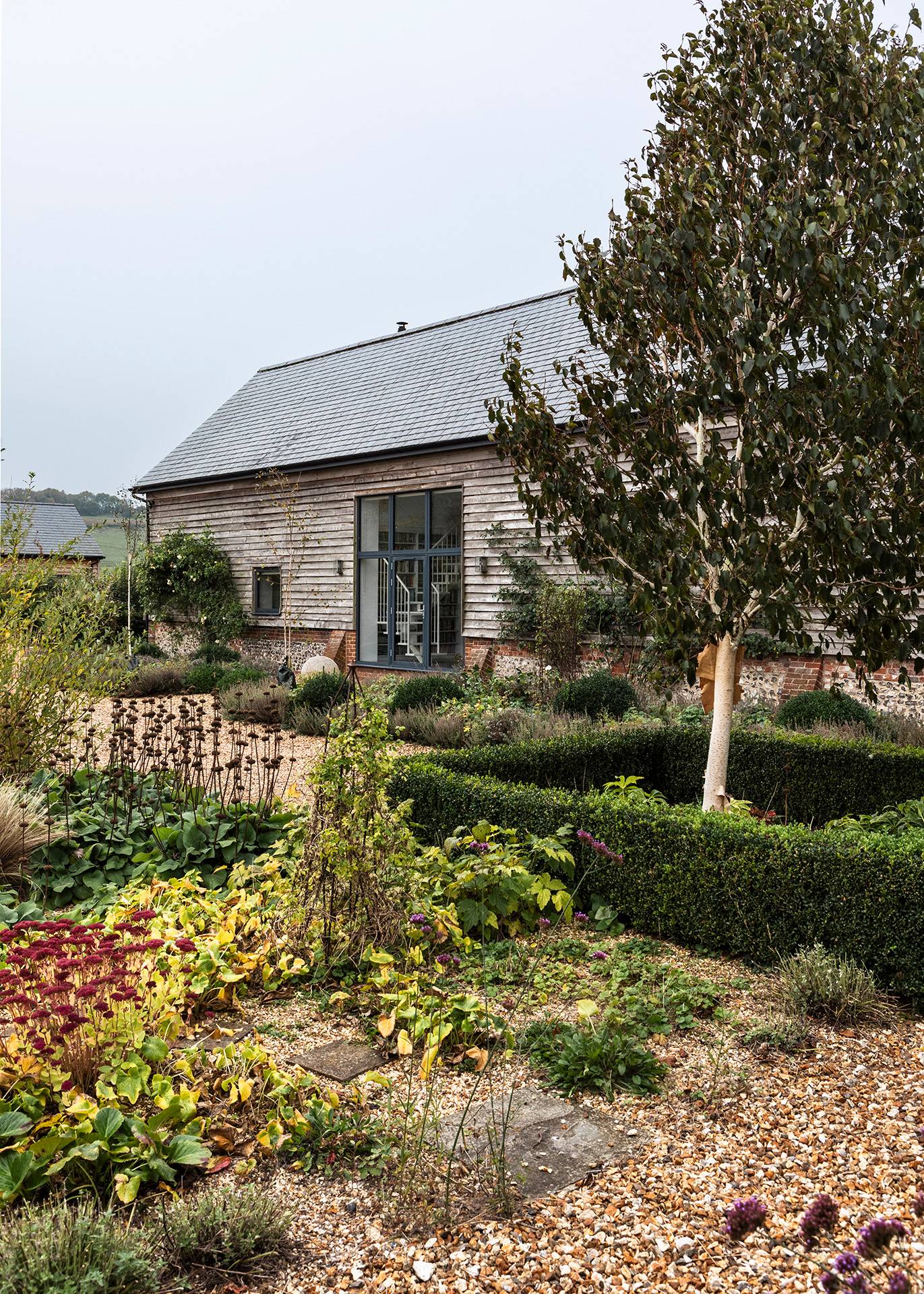 Landscaped garden beds filled with colorful perennials and boxed hedges in front of a timber barn, Love Locations SN8.