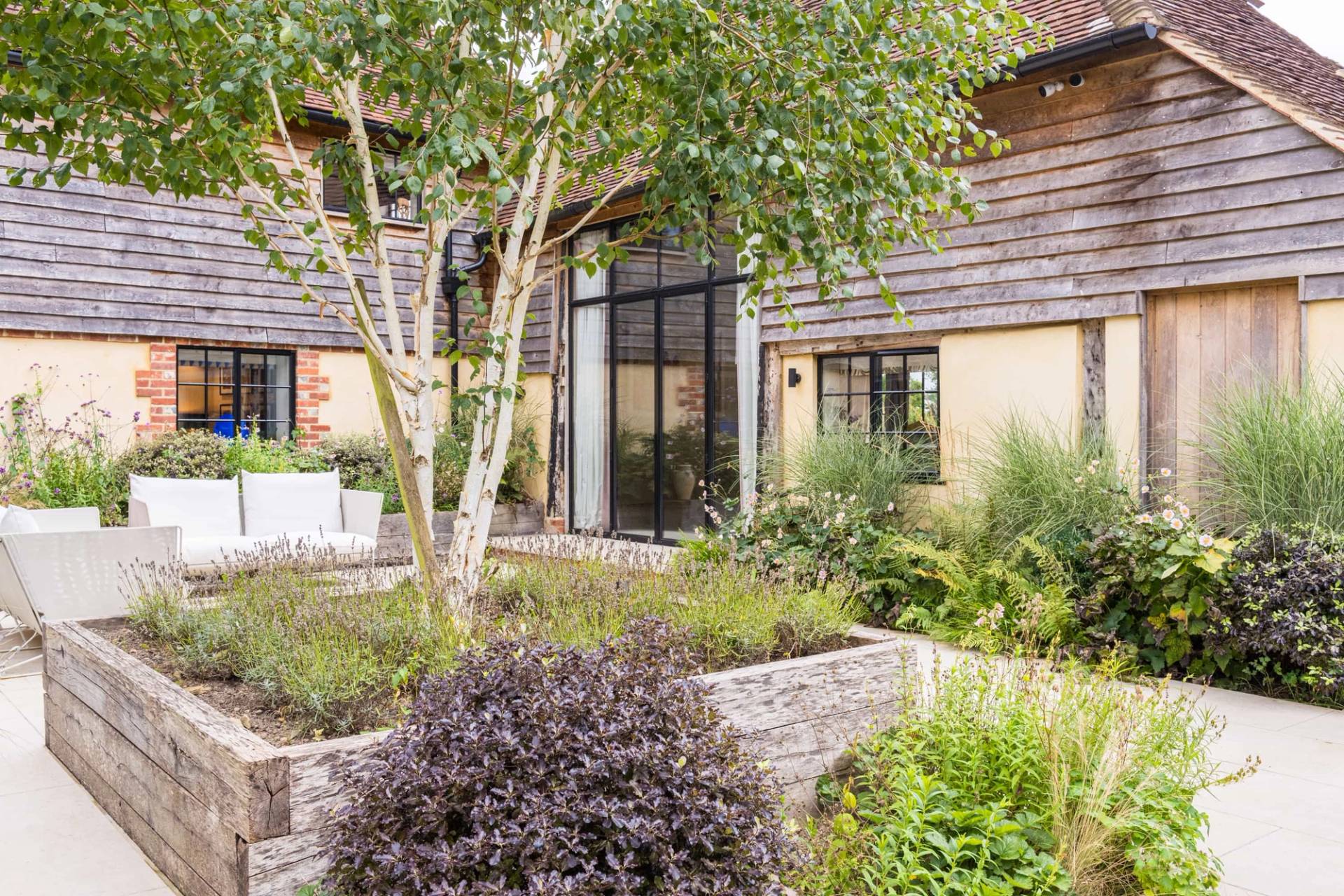 Paved outdoor terrace with courtyard gardens set outside a converted stone and timber barn house, Love Locations GU28.