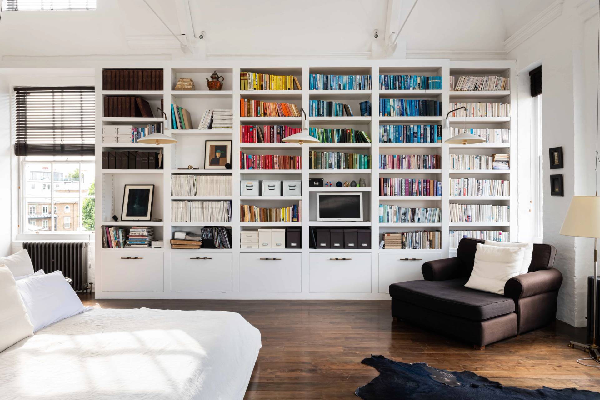 Full-wall library bookshelf setup in a white loft bedroom with dark timber flooring and plush armchairs, Love Locations.