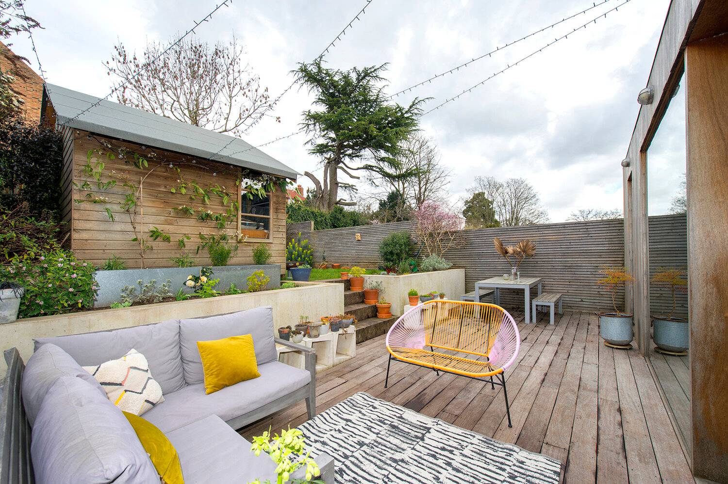 Tiered timber garden terrace featuring grey outdoor sofa seating, yellow accent cushions, and raised concrete planter beds, Love Locations SE23.