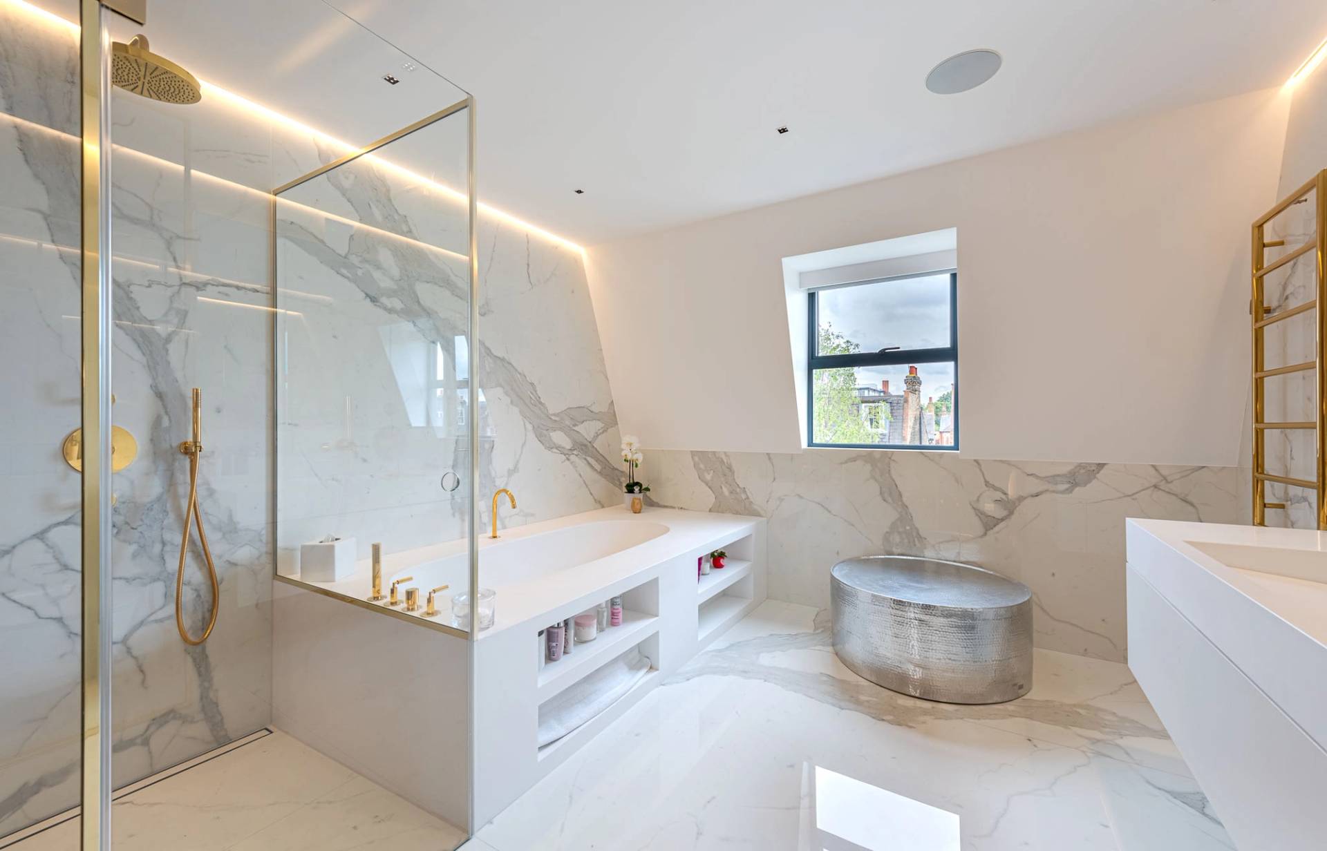 White marble bathroom suite with a glass walk-in shower, soaking tub, and illuminated niche shelves, Love Locations SW11.
