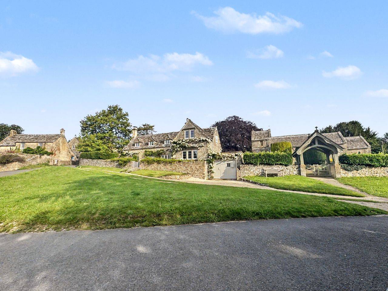 Picturesque Cotswold village street and church exterior location in Gloucestershire GL7 by Love Locations, featuring traditional honey-colored stone cottages, open lawns, and blue skies.