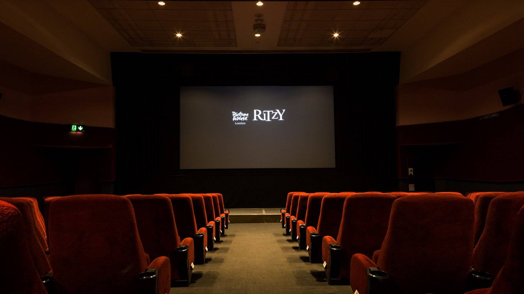 Symmetrical view down the central aisle toward the illuminated projection screen in The Ritzy Screen 2 film location in Brixton SW2, flanked by plush red velvet seats, available through Love Locations.