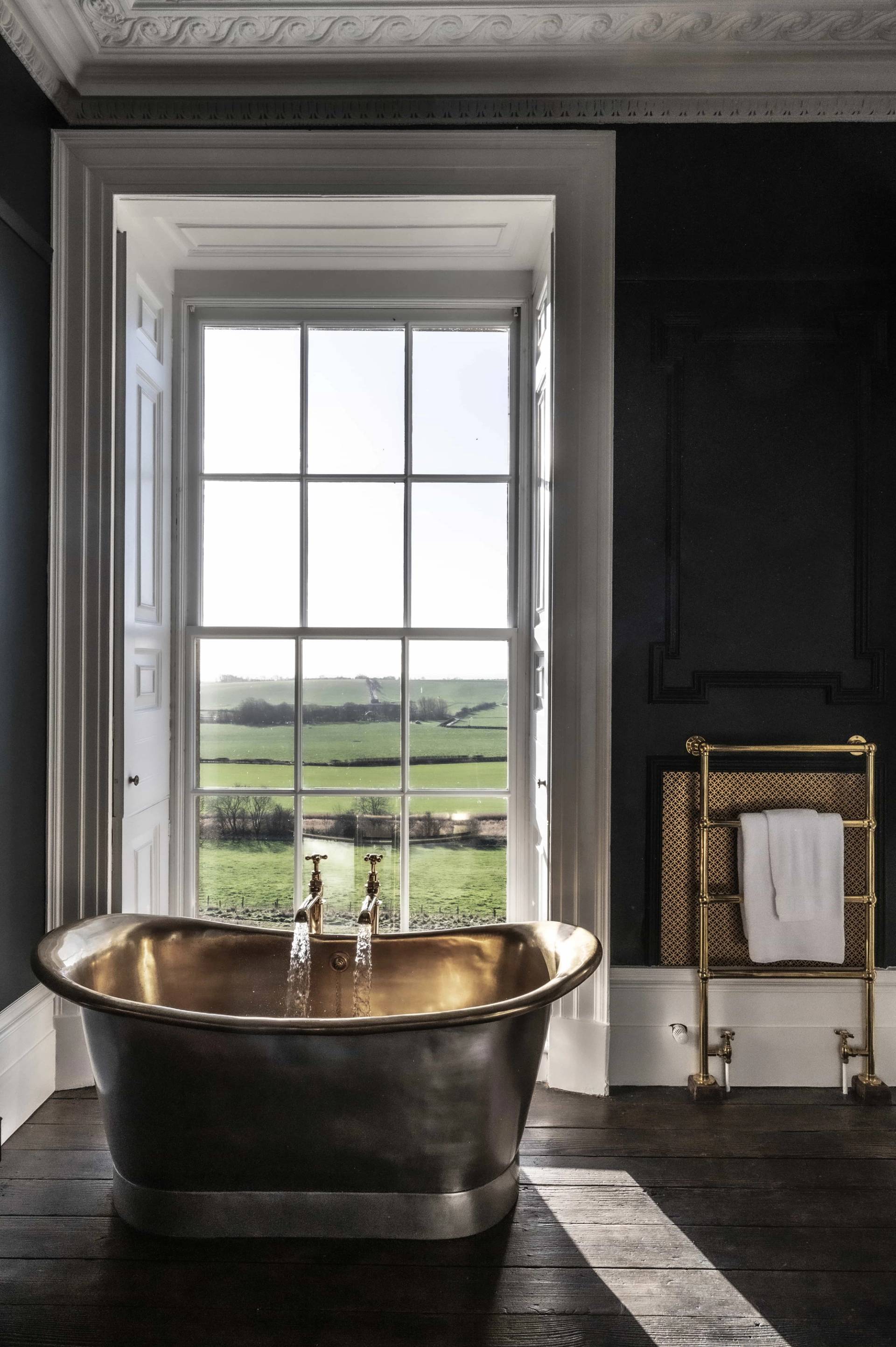 Brass clawfoot soaking tub set in front of a sash window against a moody dark green wall backdrop, ideal for wellness product shoots, Love Locations.