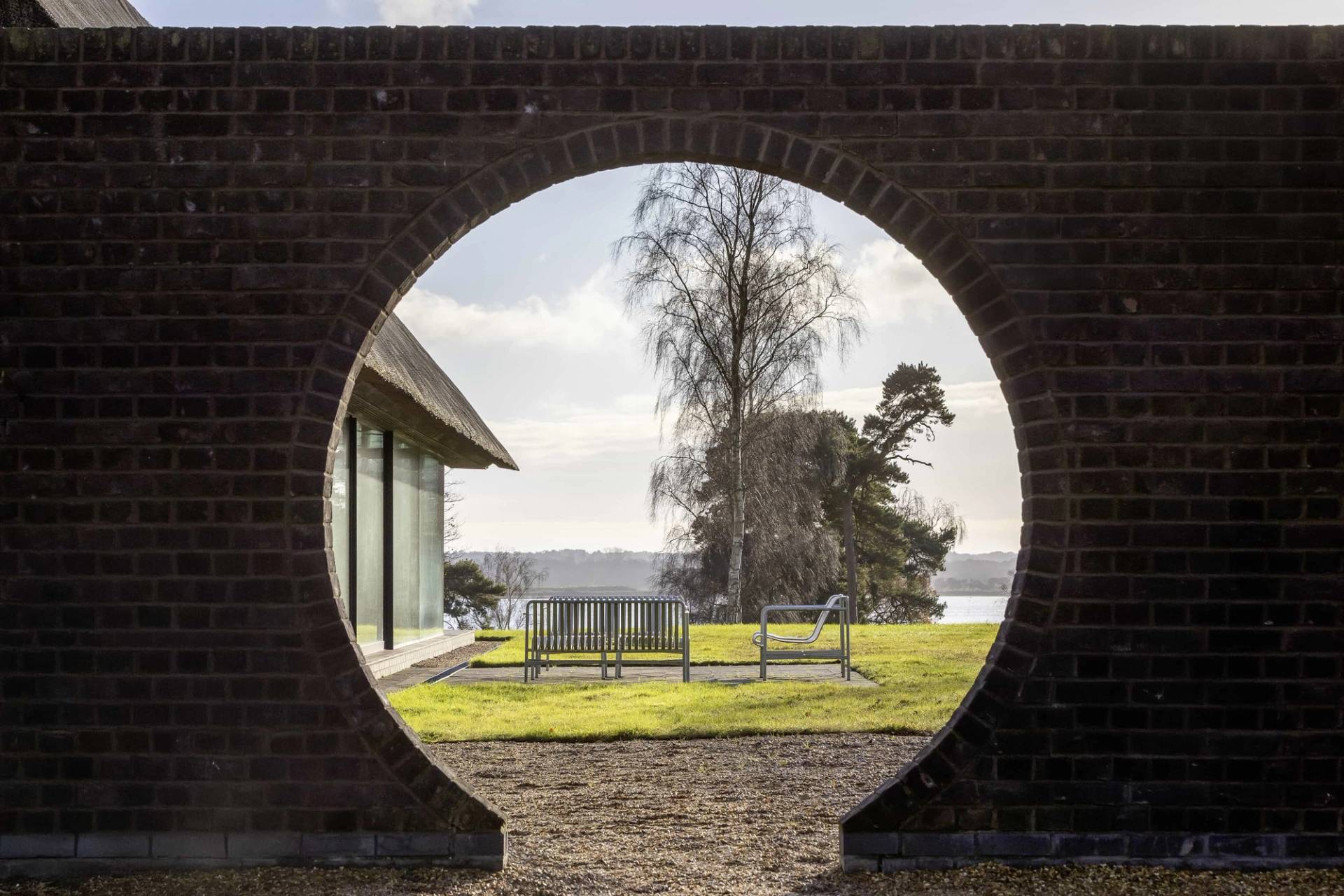 Architectural circular brick wall portal overlooking a modern thatched longhouse and rural landscape, Love Locations IP17.
