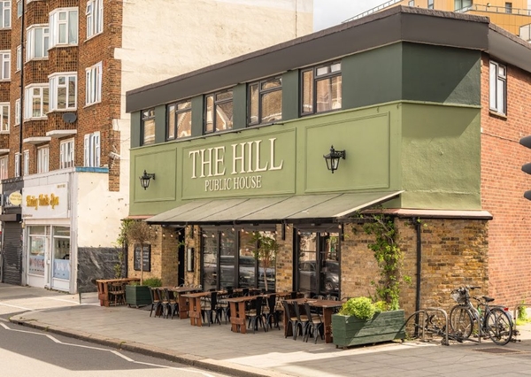 Exterior facade and building storefront establishing shot of a classic corner pub and restaurant named The Hill Public House in Streatham Hill, South West London SW16, featuring a sage green fascia, brick accents, and pavement tables, the ultimate backdrop for television drama filming and commercial production base setups via Love Locations.