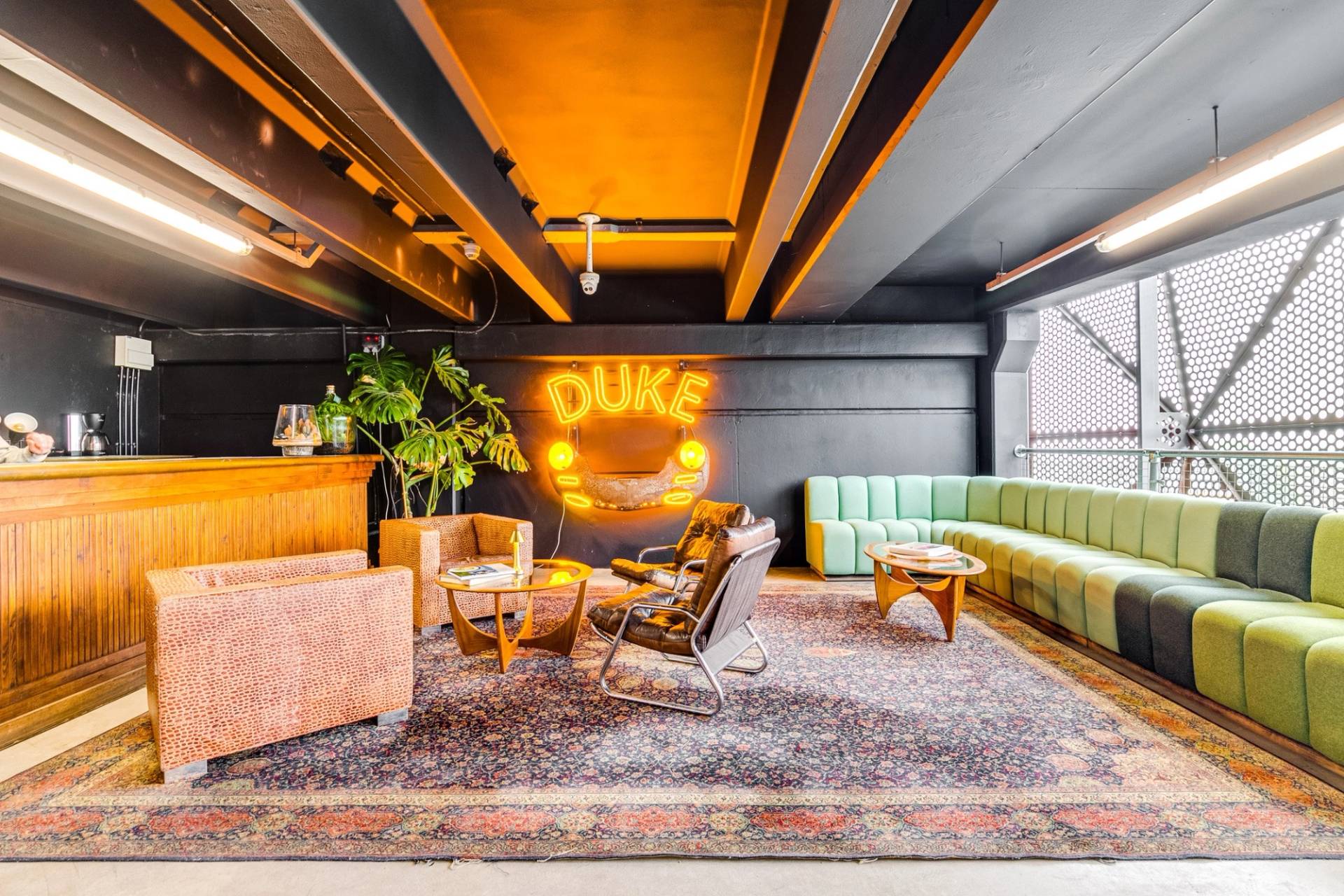 Mid-century modern lounge area shoot space in a Brentford TW8 car showroom via Love Locations, featuring a vintage Persian rug, leather armchairs, green velvet sofa, and DUKE neon wall sign.