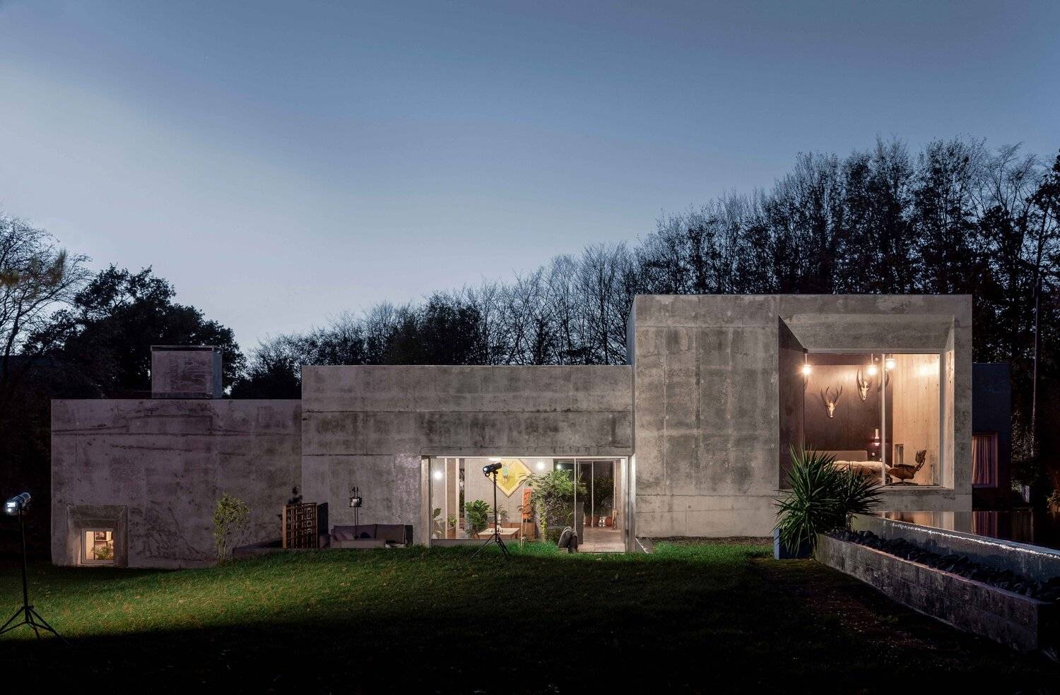 Illuminated twilight view of a brutalist concrete architectural house featuring large glass windows and lawn garden, Love Locations BN7.