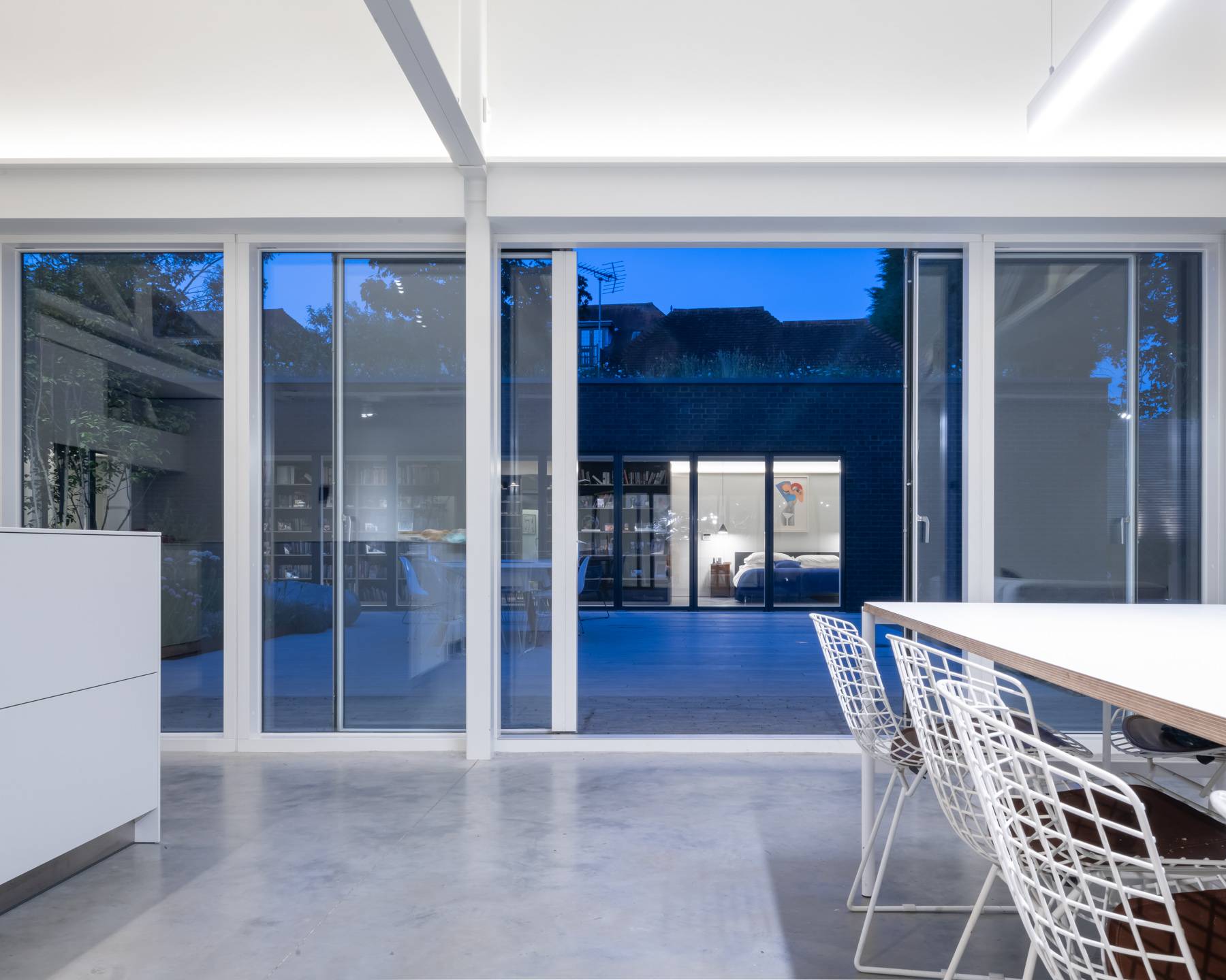 Evening perspective looking through floor-to-ceiling glass doors into a glowing white open-plan kitchen, Love Locations BN3.