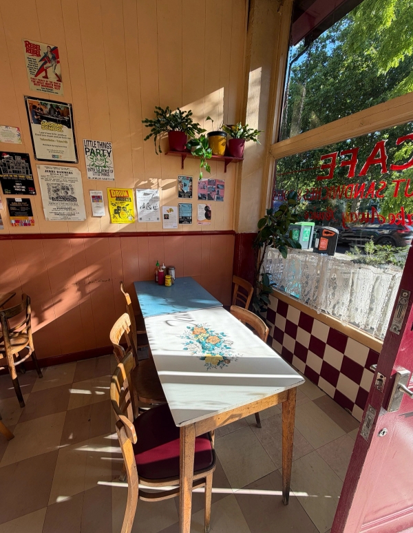 Cozy cafe window booth table with checkered wall trim, potted plants, and warm sun flare lighting, Love Locations SE27.
