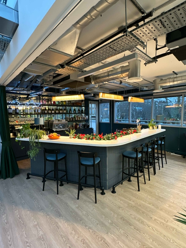 Bar area in a cool office space, featuring overhead lighting, industrial-style exposed ceiling, bar stools, and pale wood flooring