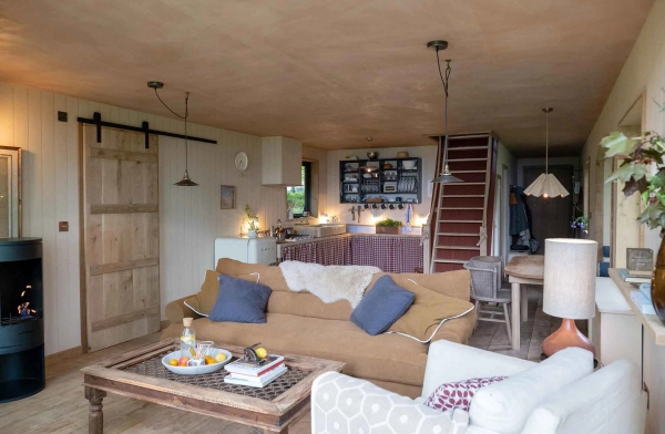Wide angle look at a cozy open-plan cabin living room featuring a tan sofa, wooden coffee table, and open stairs, great for cozy autumn film sets, Love Locations.