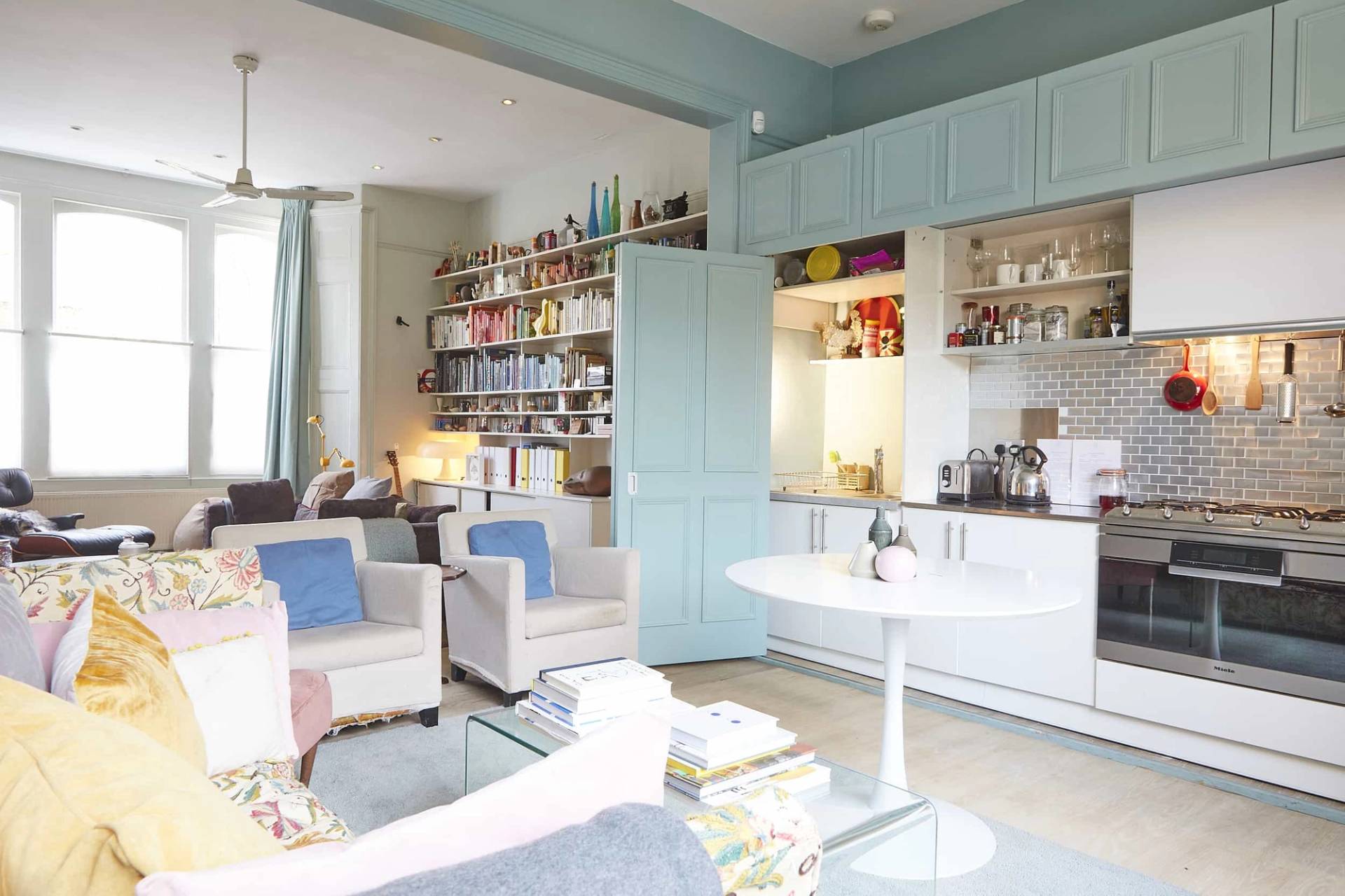 Duck-egg blue reading lounge featuring open wall-to-wall bookshelves, white armchairs, and sash window light, Love Locations N5.