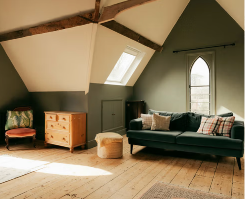 Vaulted attic room at Walton House with exposed beams, gothic-style window, painted panelling and wooden floorboards.