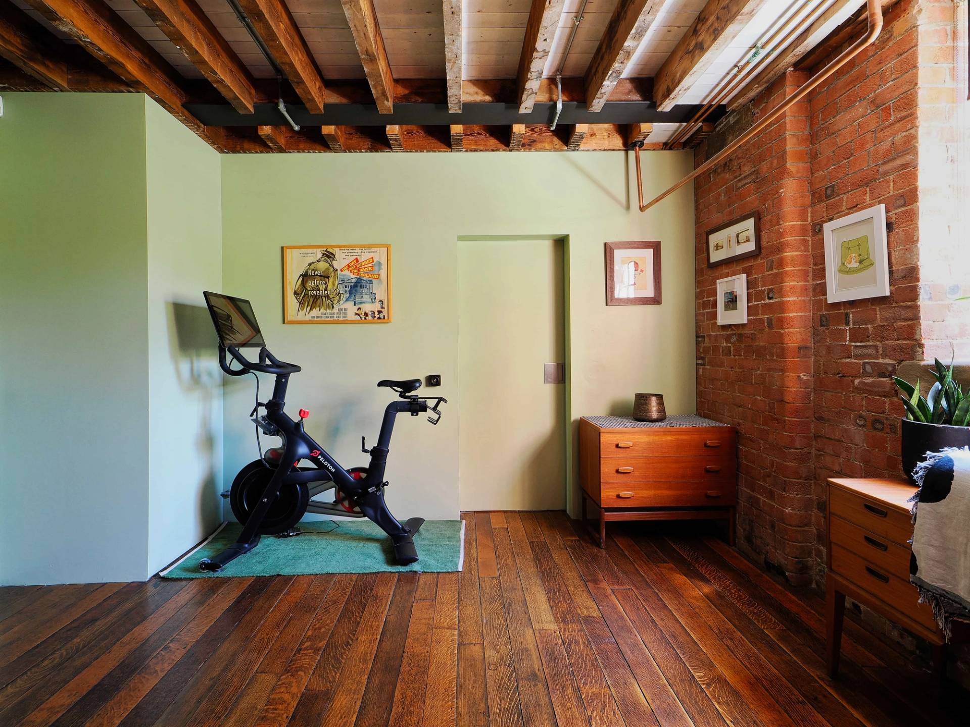 Minimalist home workout room with Peloton exercise bike, dark timber floors, and exposed brick details, Love Locations E14.
