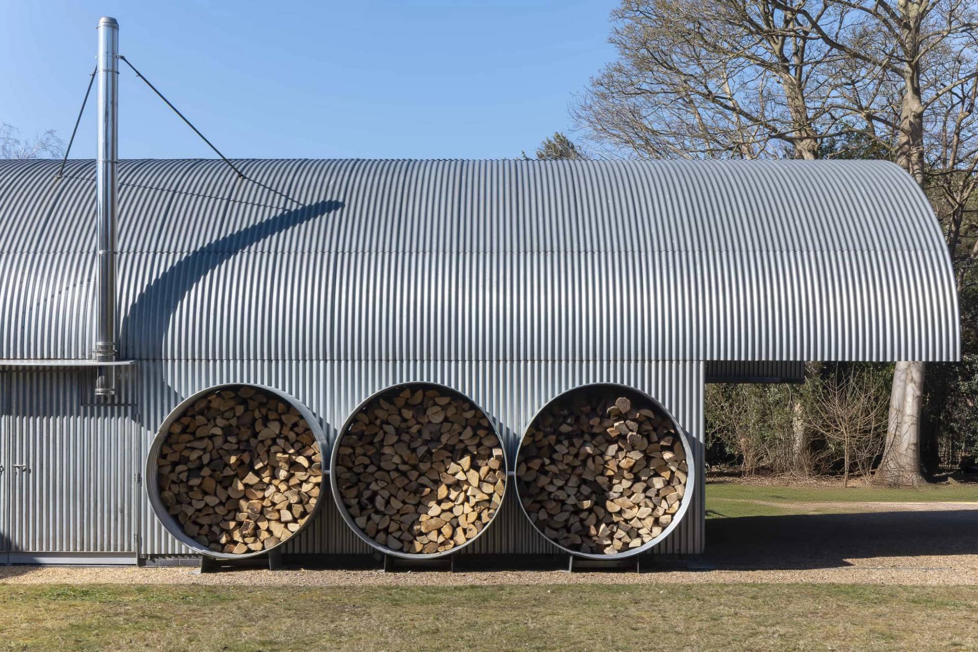 Curved corrugated metal outbuilding with integrated circular log storage alcoves filled with stacked firewood, Love Locations IP17.
